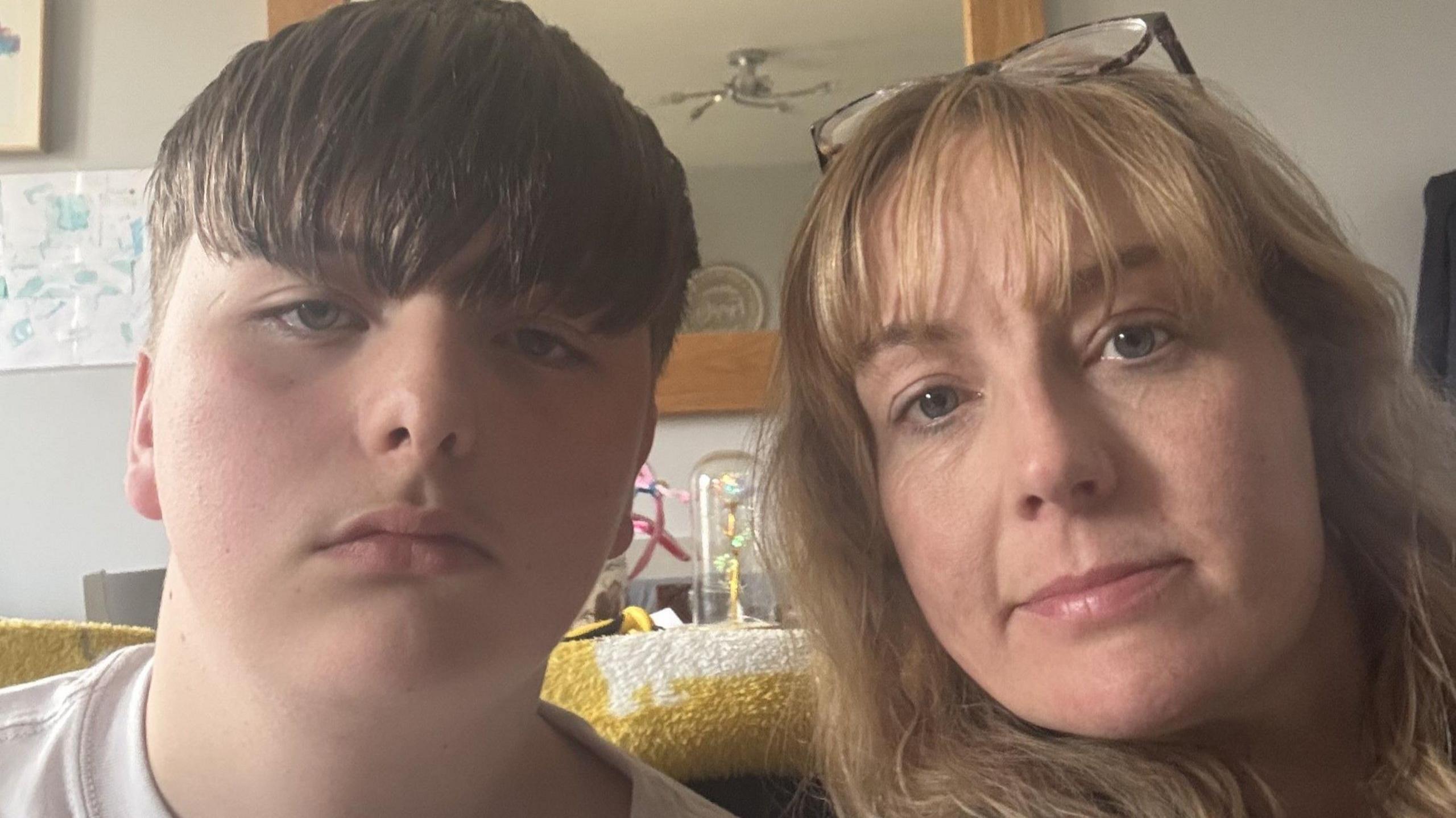A boy with short brown hair takes a selfie with a woman with long, blonde hair and glasses on her head. The pair are sat in a living room setting with a mirror behind them.