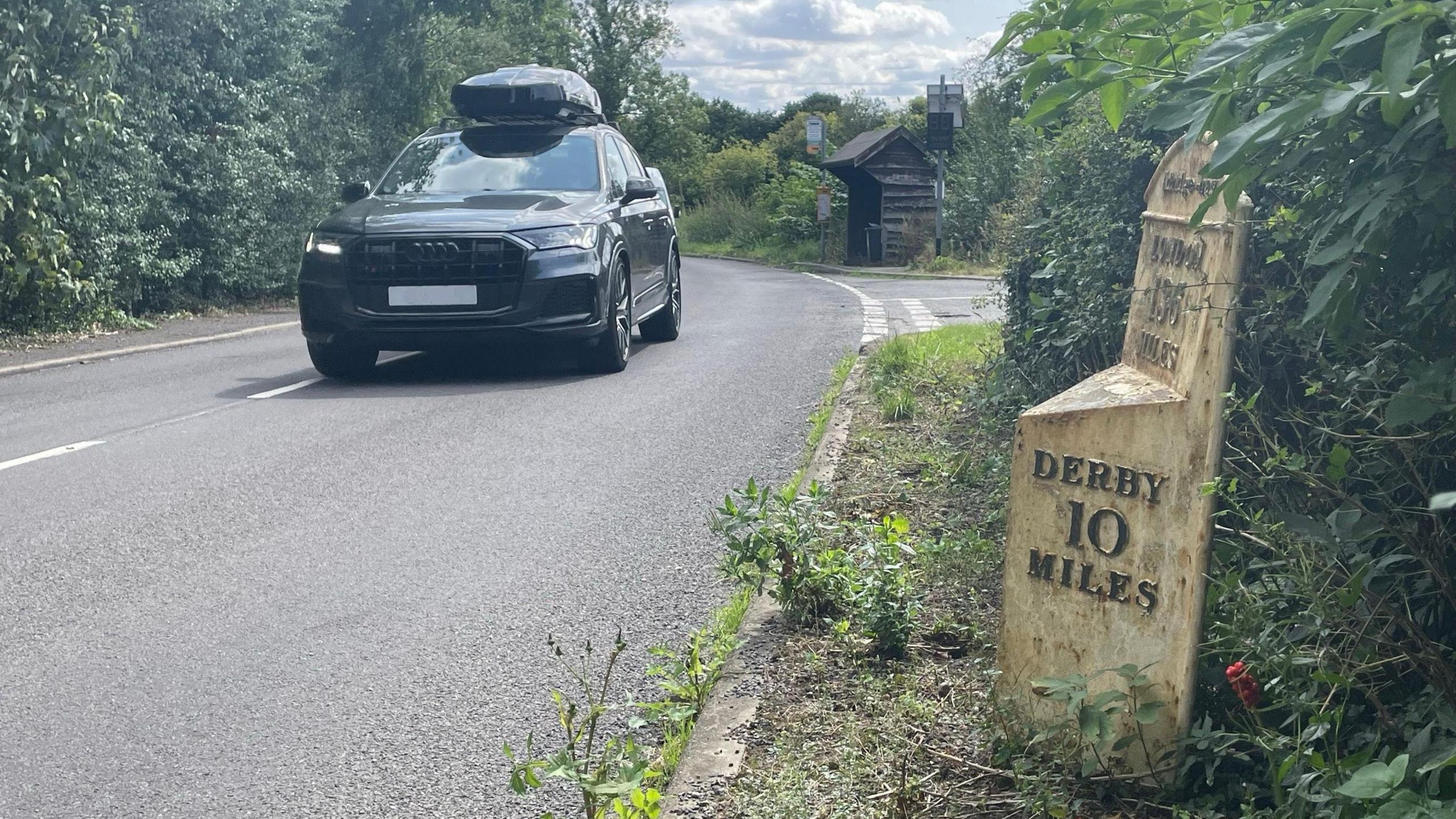 A cast iron milestone by the side of a road in a village with a car driving past