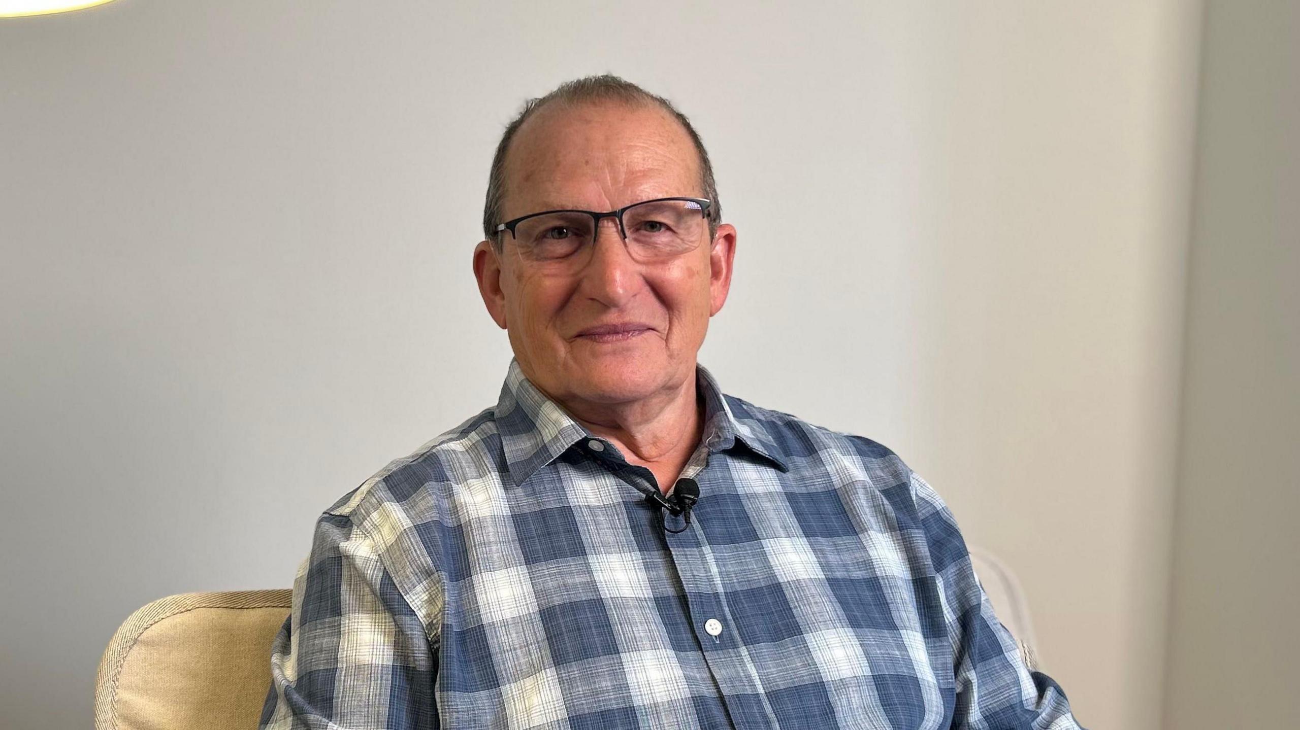 Don Connolly in a chequered shirt sits on a chair. He is wearing glasses and smiling at the camera.