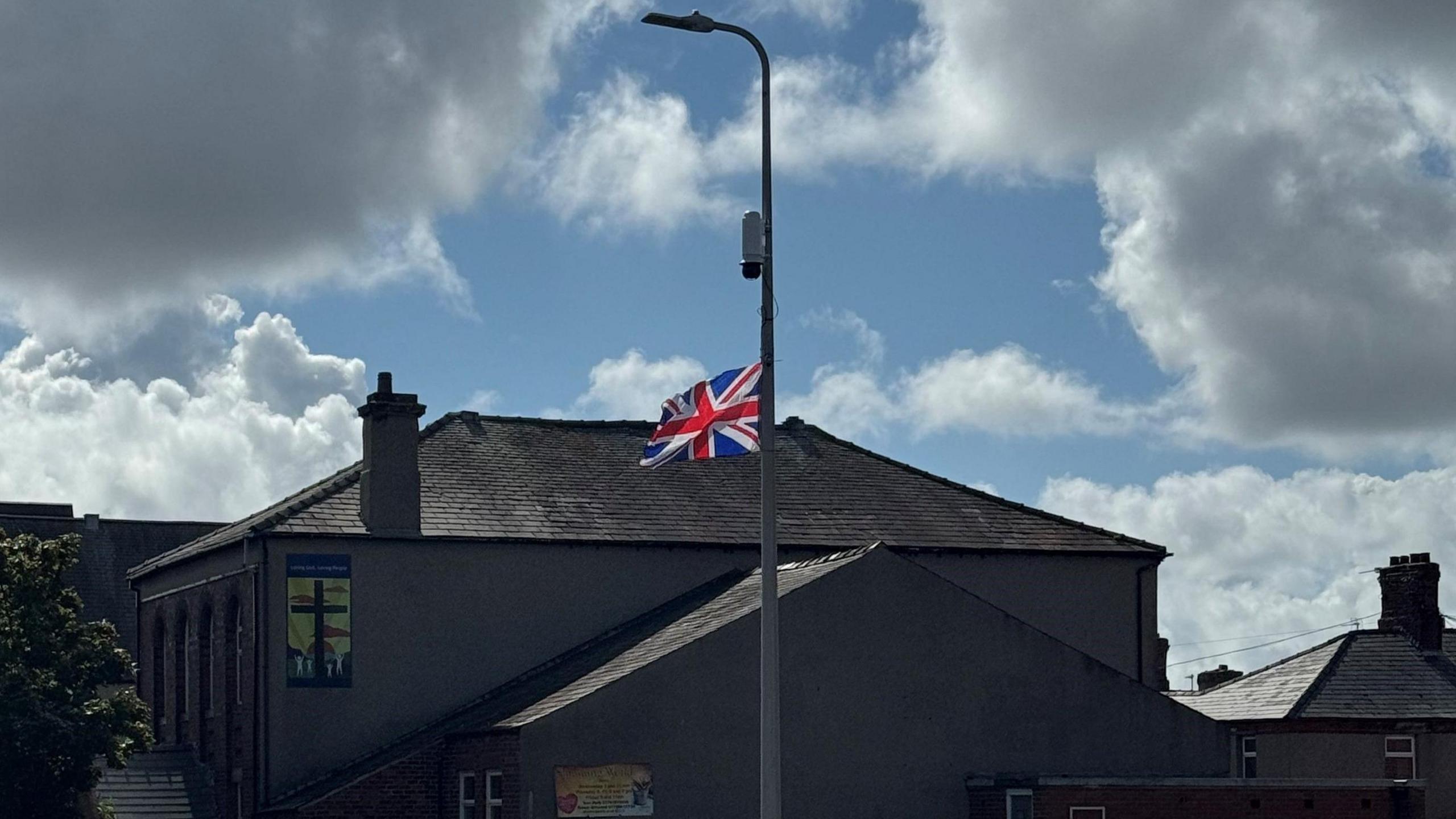 A picture of the lamppost with the union jack flag attached. The flag is billowing in the wind about half way up the lamppost. It has been hung upside down. Above it, a CCTV camera has been attached to the lamppost. A church stands behind the lamppost and has a poster displaying a cross on its wall.