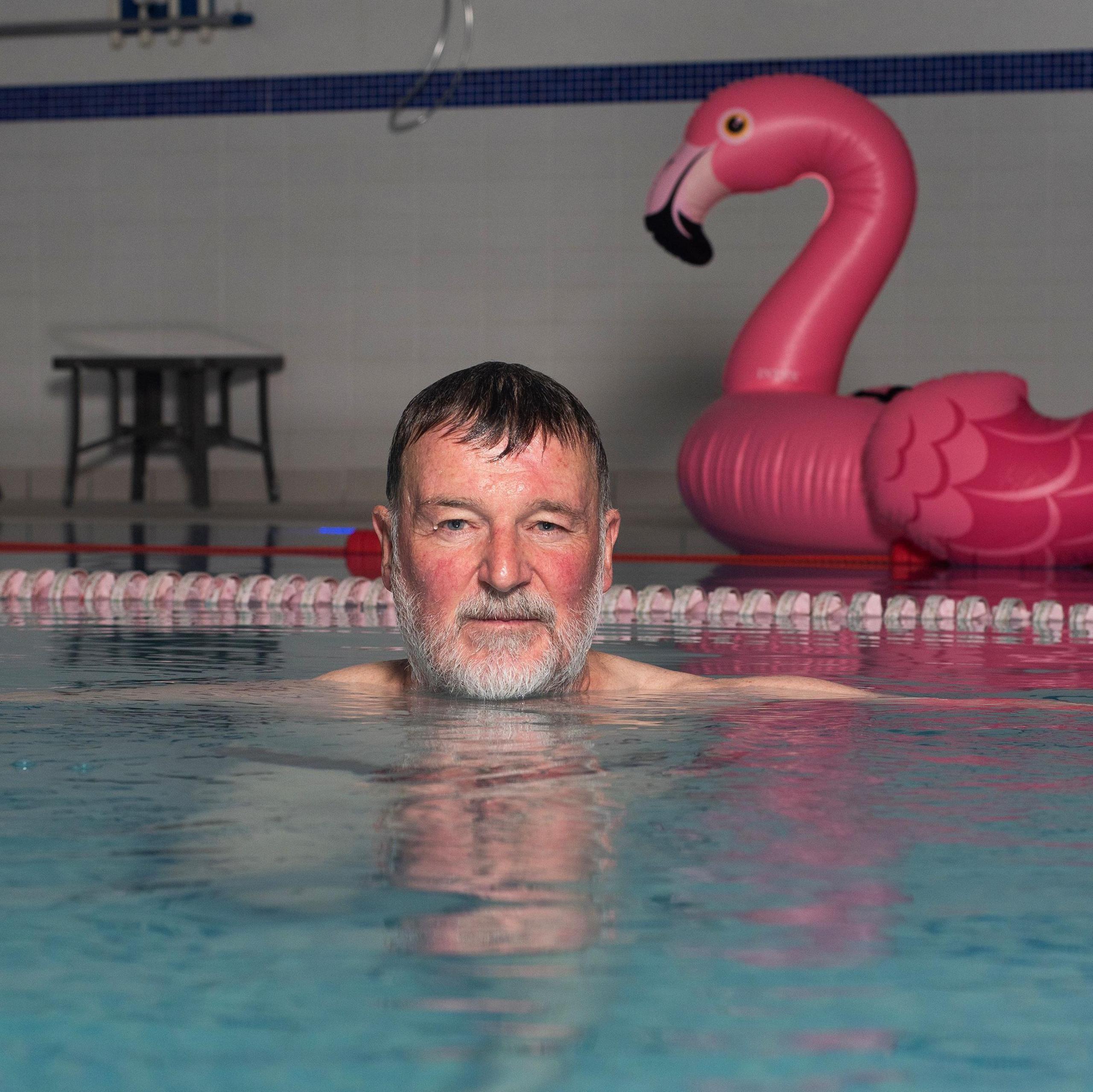 Man looking in the camera in a swimming pool with a his head visible above the water. A plastic flamingo is in the background. 