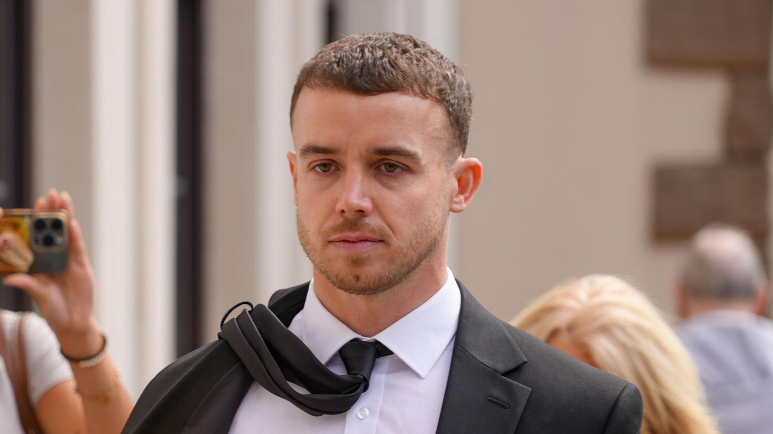 Lewis Carr walks to court with a black suit and white shirt. He has a black tie on which is slung over his right shoulder.