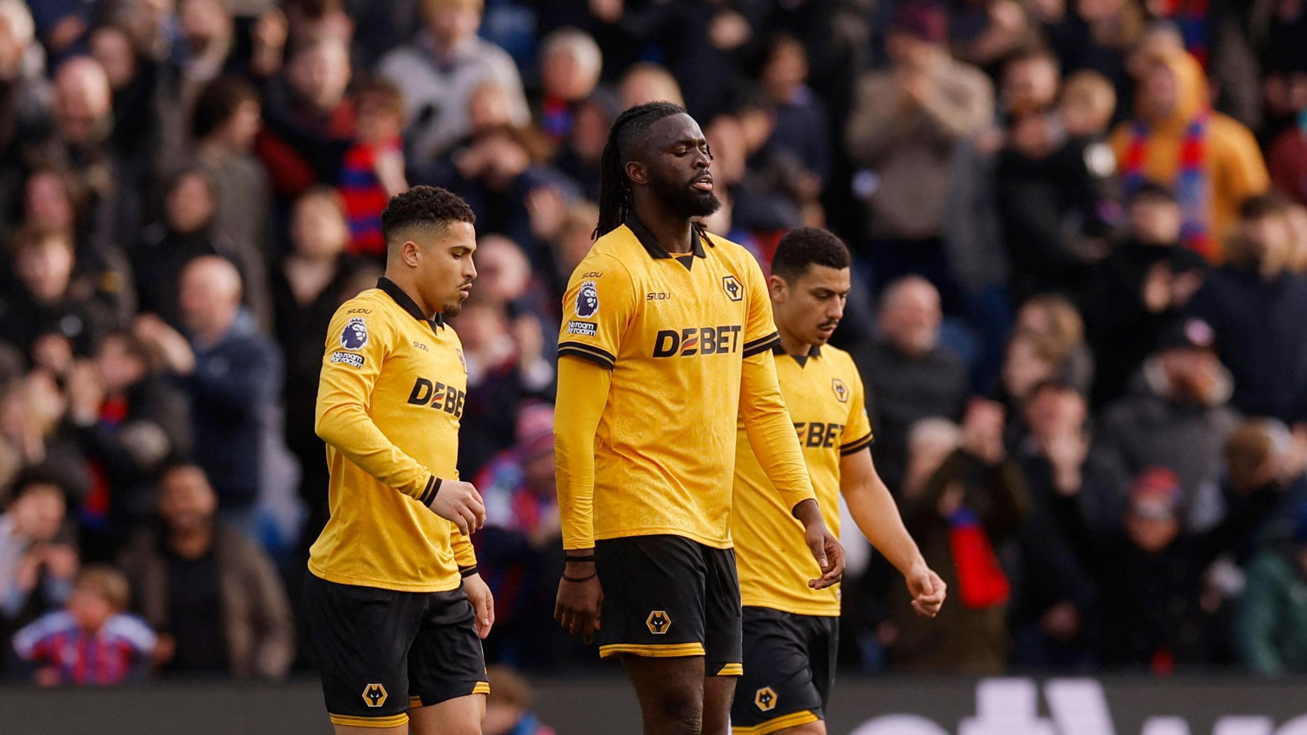 Wolverhampton Wanderers' Tolu Arokodare wears a gold and black football kit and is walking across a pitch. There are two other players either side of him and crowds blurred in the stands behind. He has his eyes closed as he walks and looks dejected.