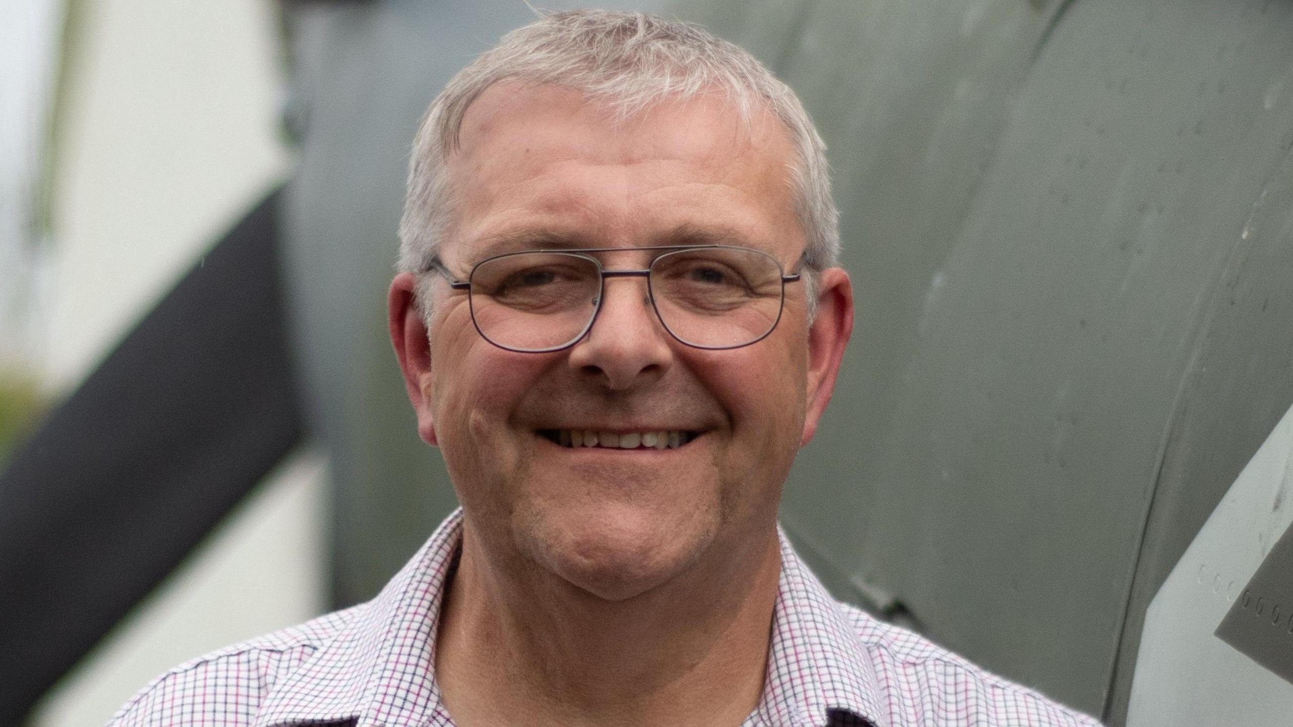 David Price standing in front of the Spitfire he built. He has short white hair and glasses and is wearing a shirt with a chequered pattern. Behind him is the spitfire is painted a brown-grey colour.