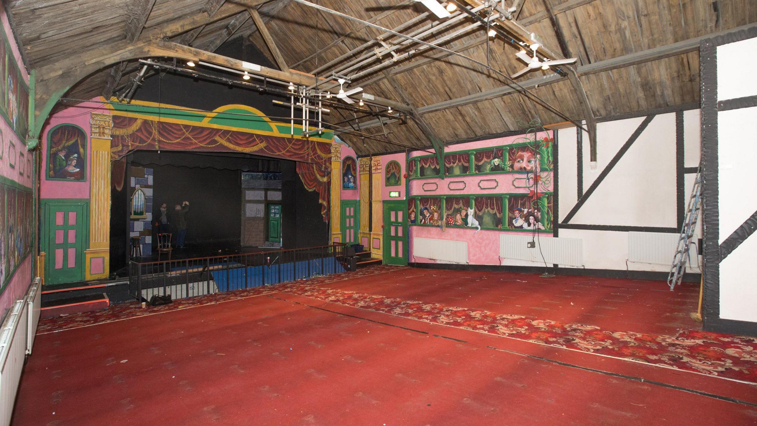 An inside view of the theatre, it has a wooden panelled A-shape ceiling with lighting attached to a central panel. There is red carpeting on the floor and the walls are a mixture of pink and green paintings for plays and black wooden beaming with white walls. There is a stage with a black rail around the front.