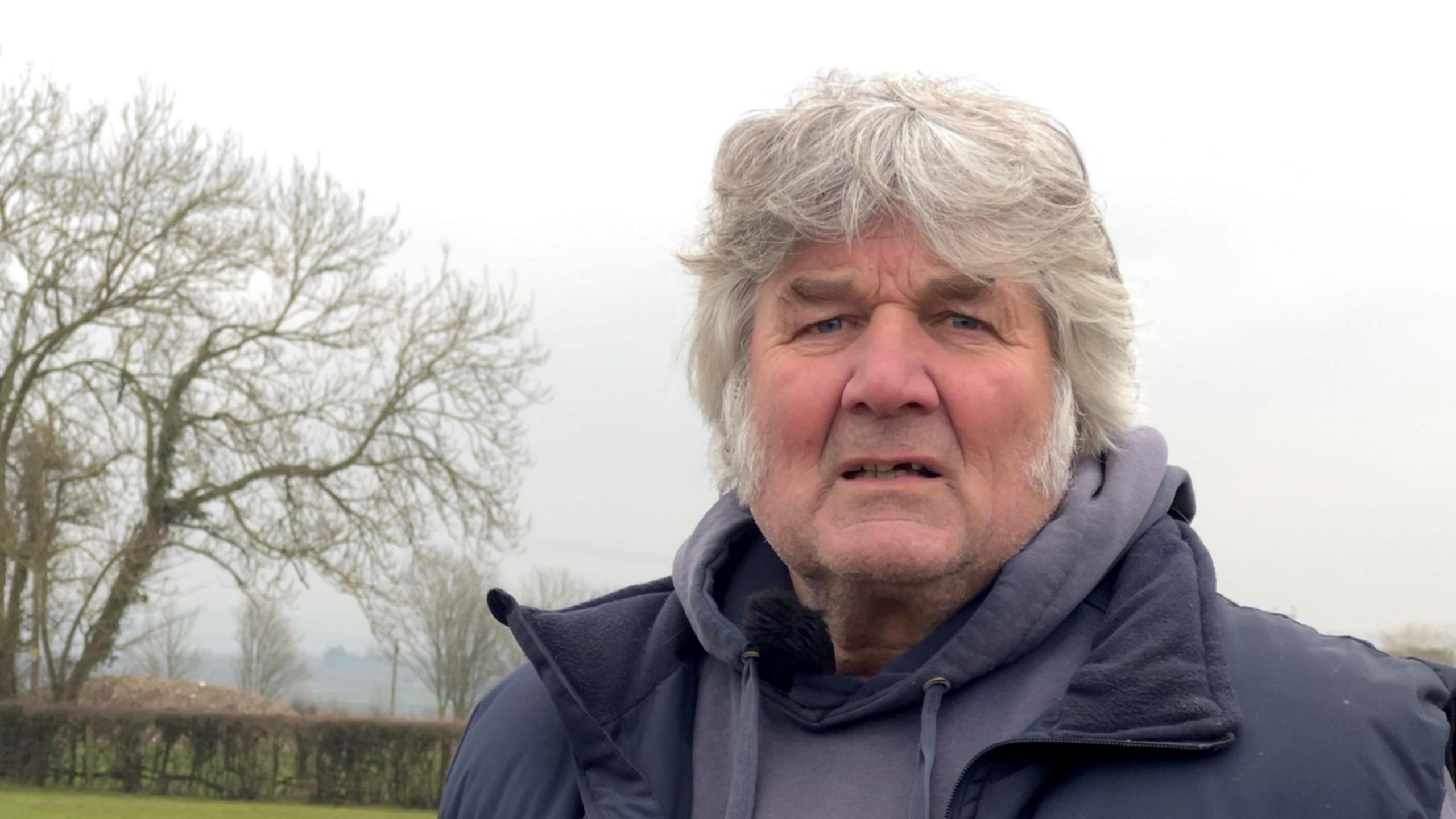 Man with grey hair looking at the camera whilst stood on a farm. There's a tree in the background.