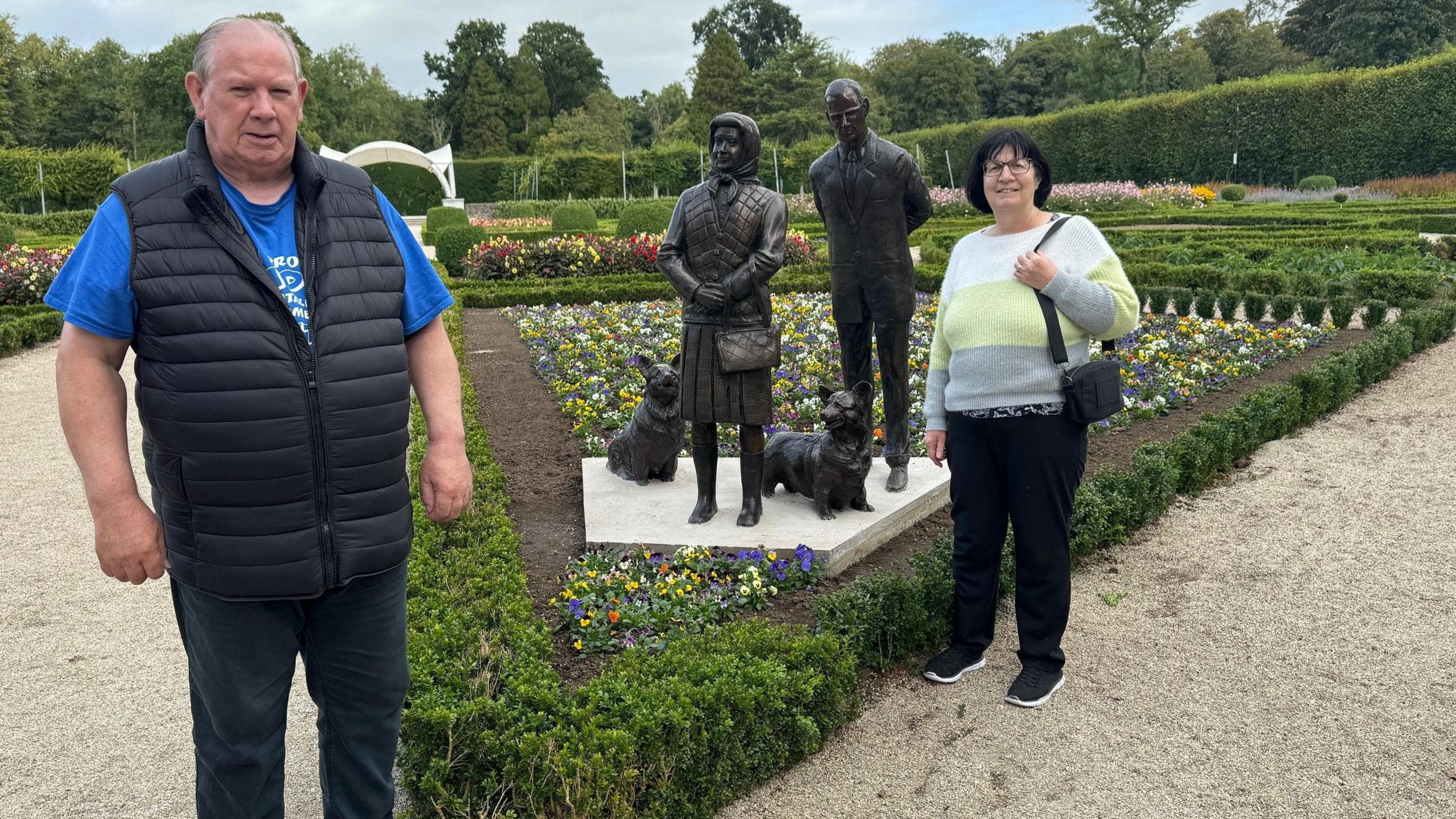 Francis and Marie are standing on opposite sides of the sculptures. 

Francis is tall, has grey short hair and is wearing a black puffer jacket over a blue t-shirt. He is wearing dark denim jeans. He is smiling. 

Marie has jet black hair in a bob cut. She is wearing glasses and is carrying a black handbag. Her jumper is white, yellow and grey and her shoes and trousers are black. She is smiling. 
