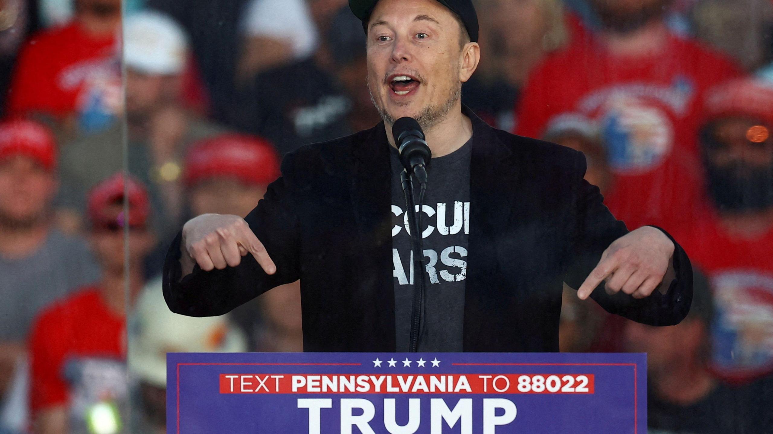 Elon Musk, wearing a black 'Make America Great Again' cap, a black blazer and grey emblazoned t-shirt, points to a sign for Donald Trump's presidential campaign as he speaks at a Trump rally in Pennsylvania 