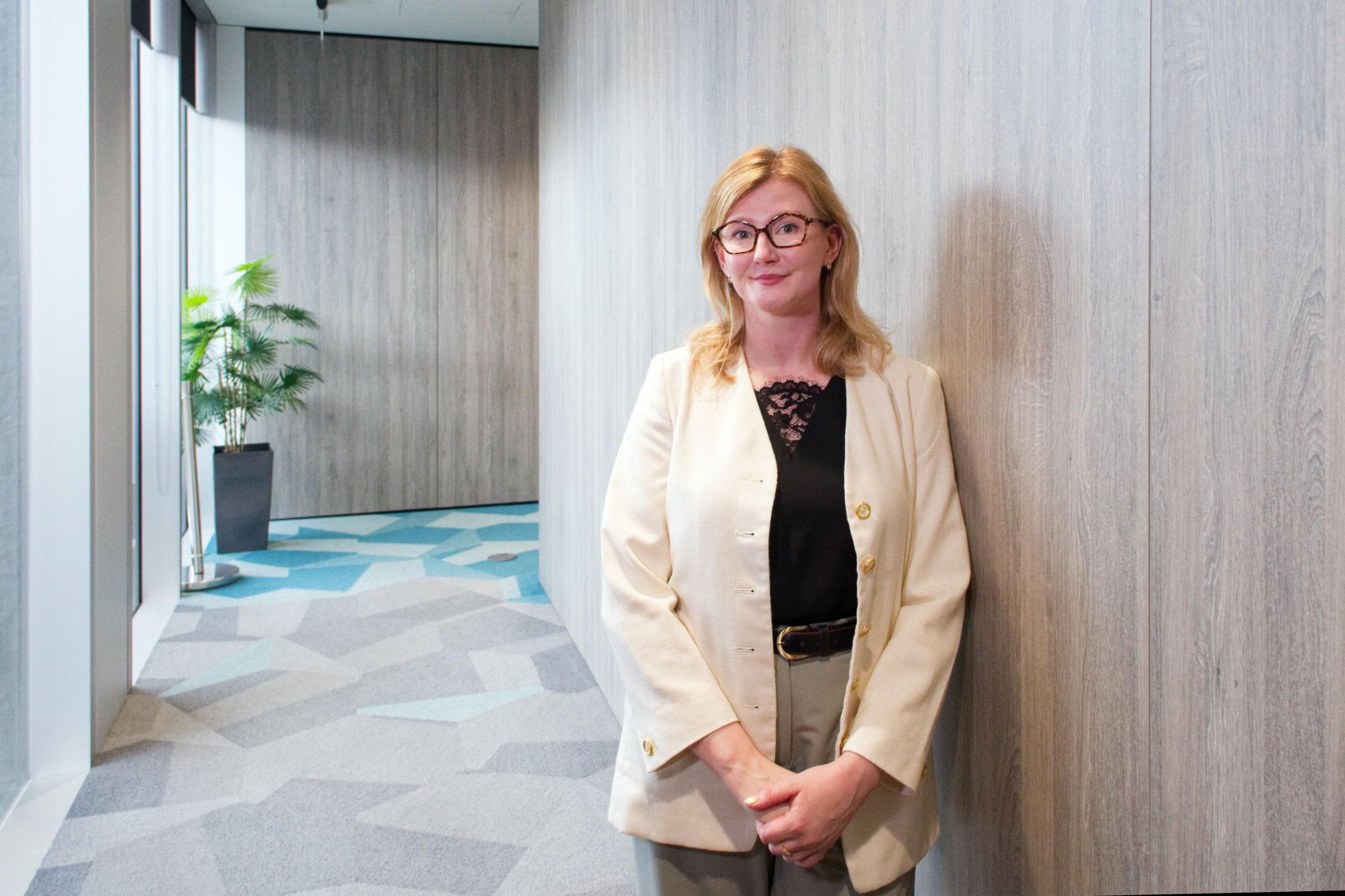 Karina Hutchins has her hands clasped in front of her in a light-filled corridor at UK Finance's HQ building in central London. Behind her is potted palm-type plant. Karina wears glasses and has stawberry blonde hair. She is smiling. She is wearing a cream-coloured business jacket and trousers and a black blouse beneath.