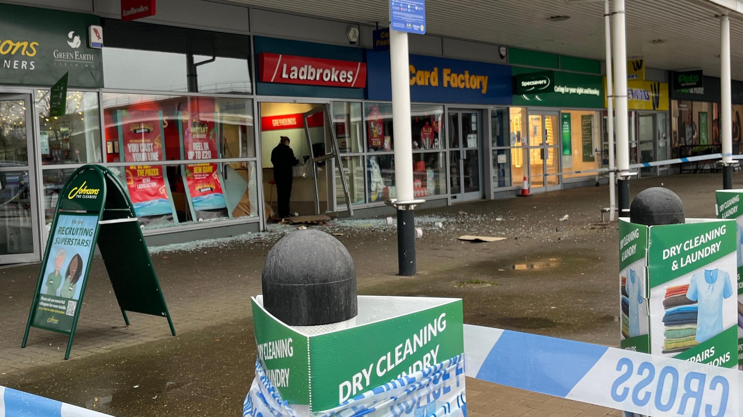 A row of shops under white awning held up by pillars. The one in the middle has Ladbrokes written in white letters on a red background above its door. The door his on its hinges, the glass smashed and in piles on the ground. A person in a boiler suit can be seen at the count of the building. In the foreground is blue and white police tape. 