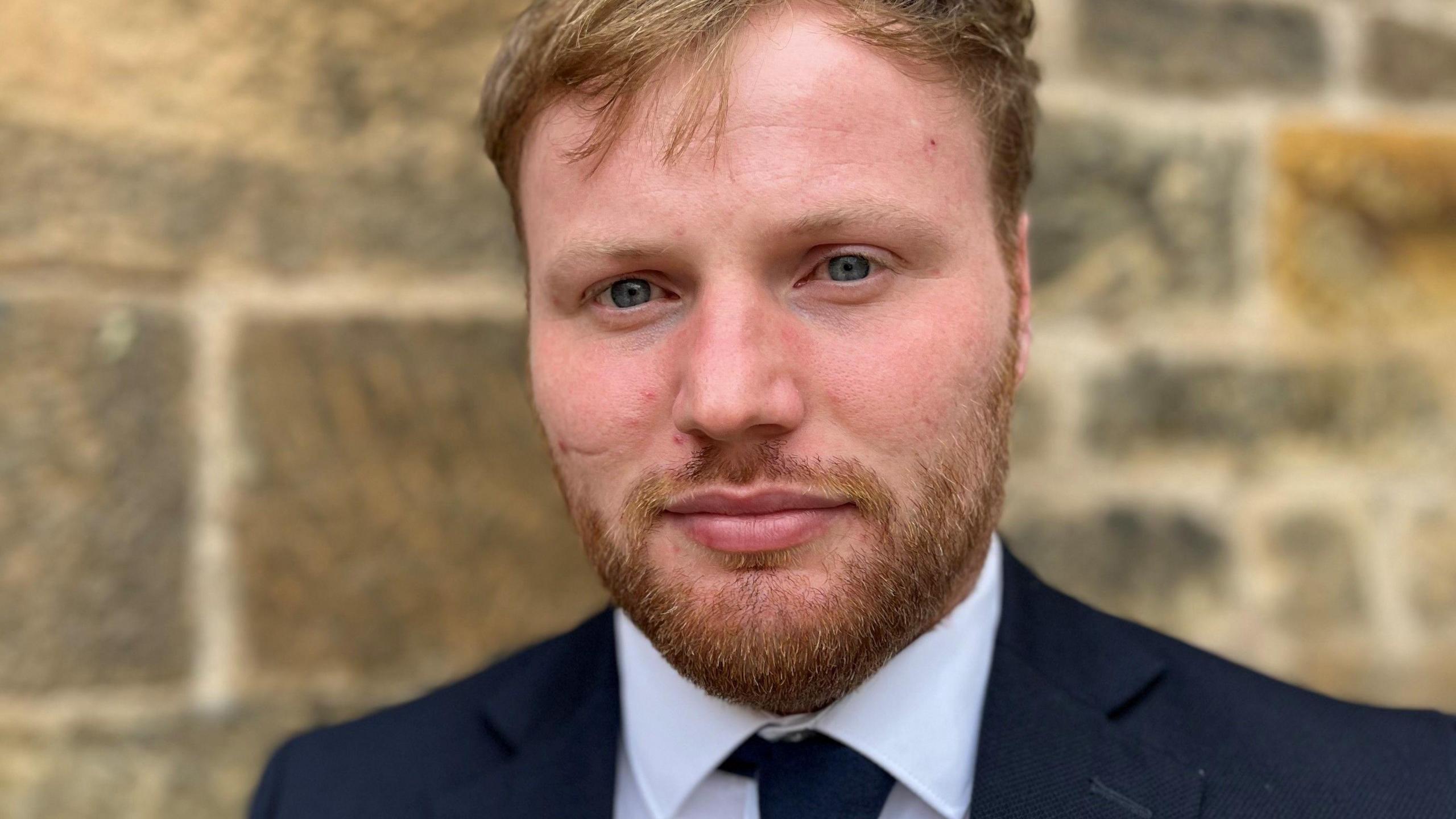 Lawyer Andrew Fyffe is photographed outdoors against a stone wall. He has shoirt hair and neatly trimmed ginger beard. He is wearing a dark suit, white shirt and blue tie