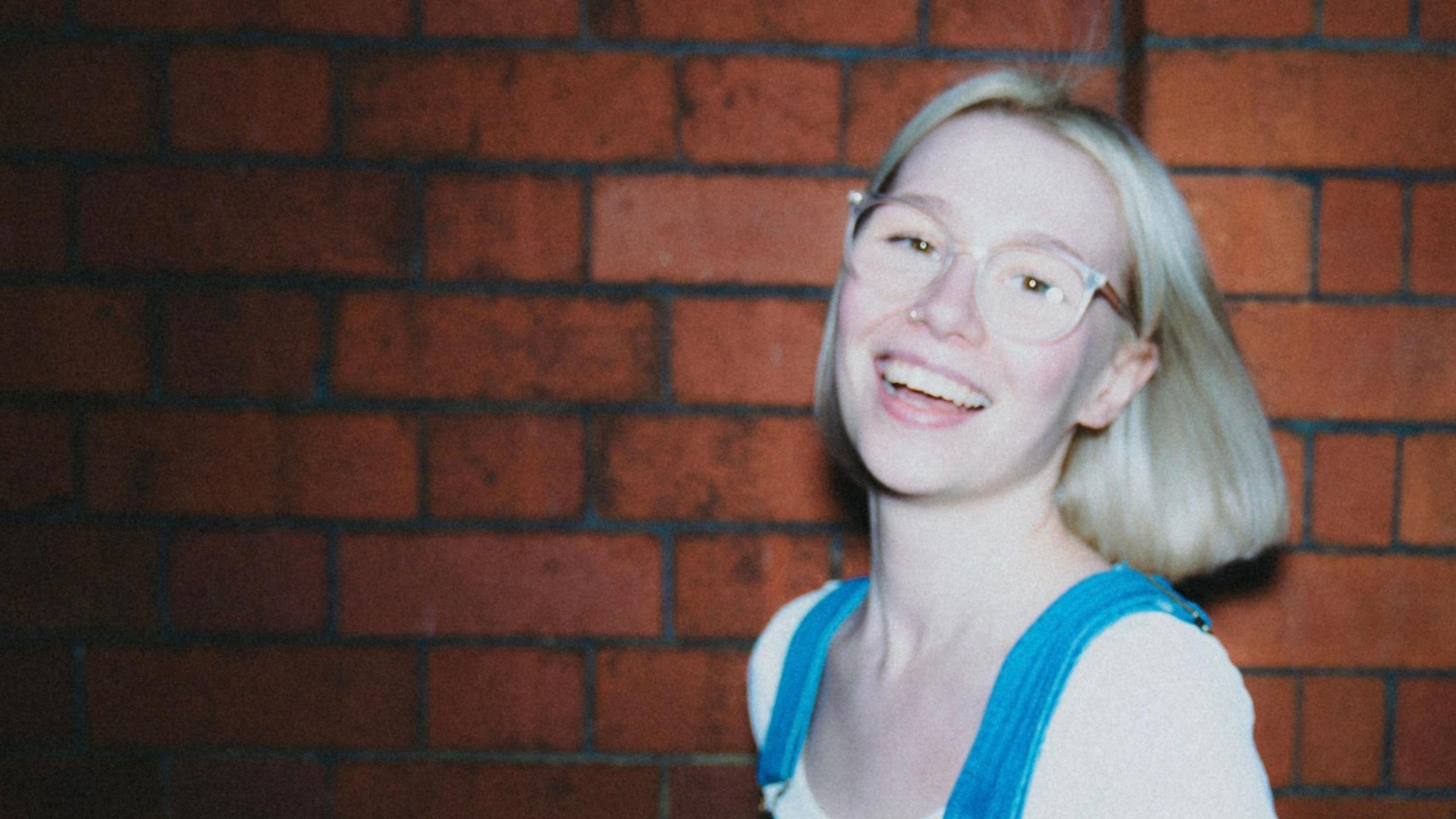 Molly Palmer, who has blonde bobbed hair and is wearing a blue pinafore and white t shirt, smiling at the camera with a red brick wall behind her