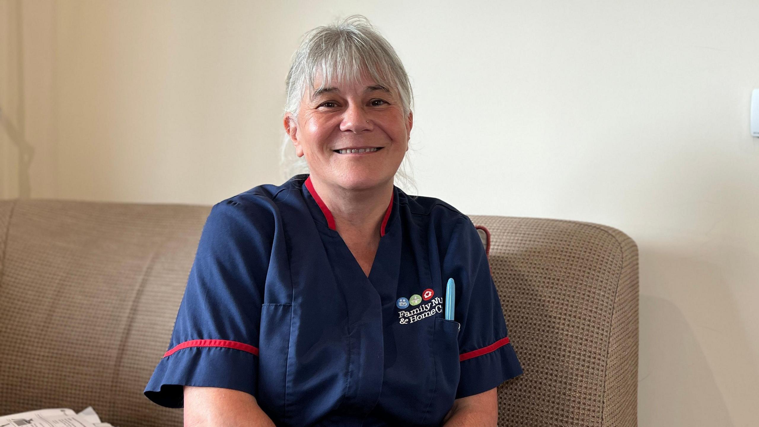 Gilly sits on a grey sofa in her uniform and smiles at the camera