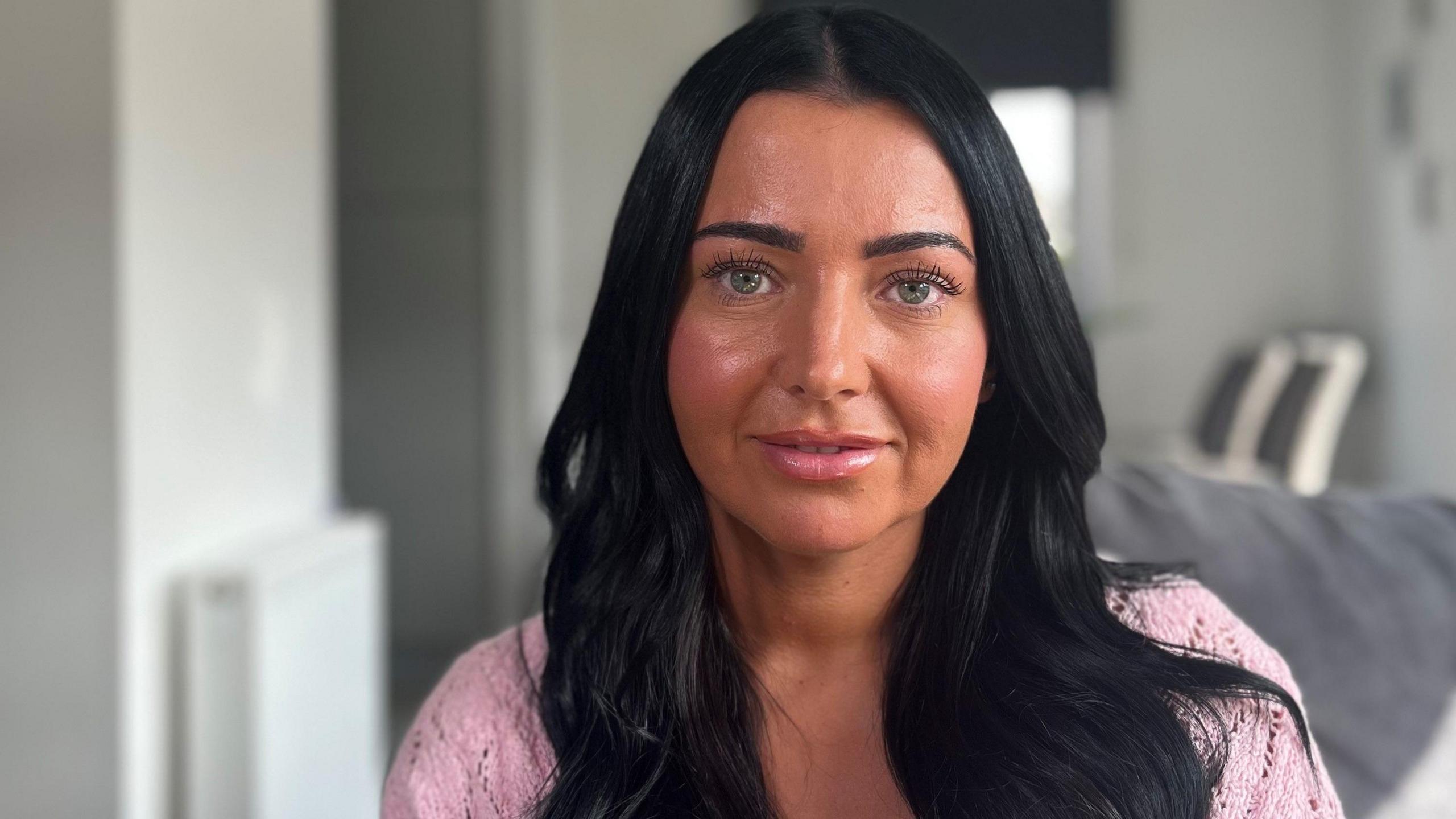 Mum Samantha is photographed indoors. The background is blurred. She is looking directly at the camera. She has long black hair and is wearing a pink top