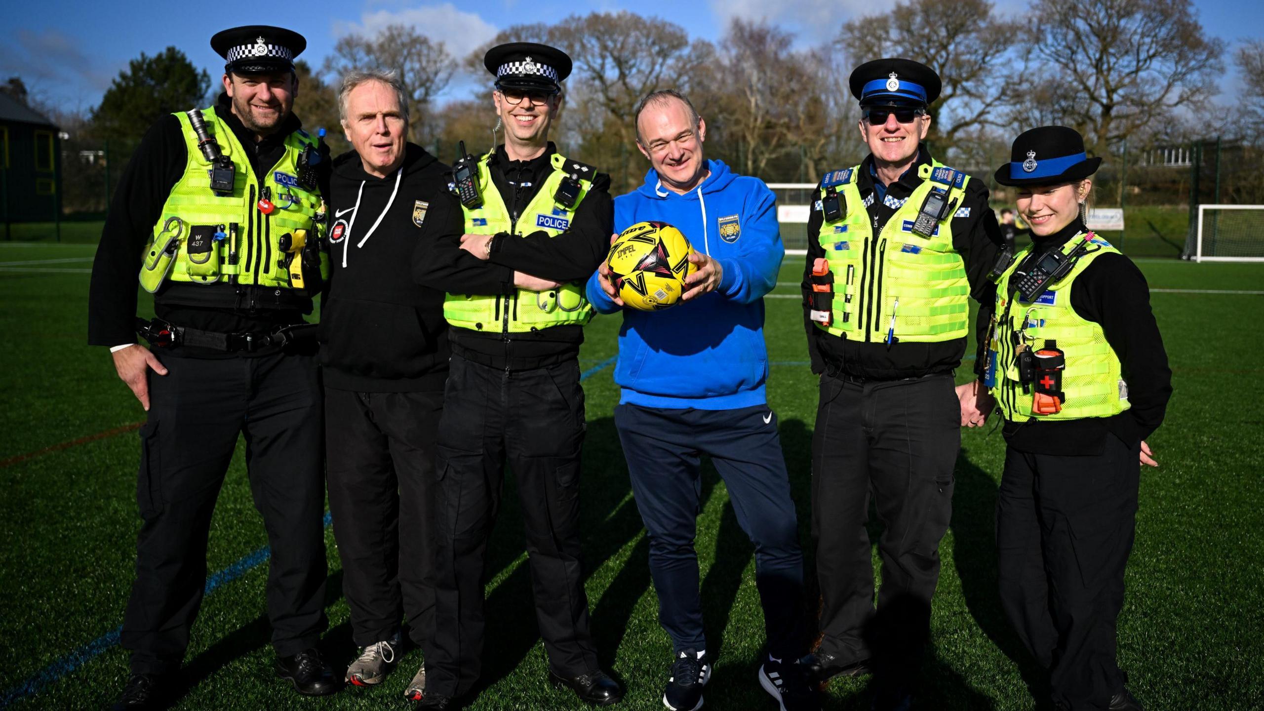 Liberal Democrat leader Ed Davey stands alongside a group of police officers in uniform. He is holding out a yellow football and they are all stood on a football pitch at Wiltshire FA headquarters in Devizes