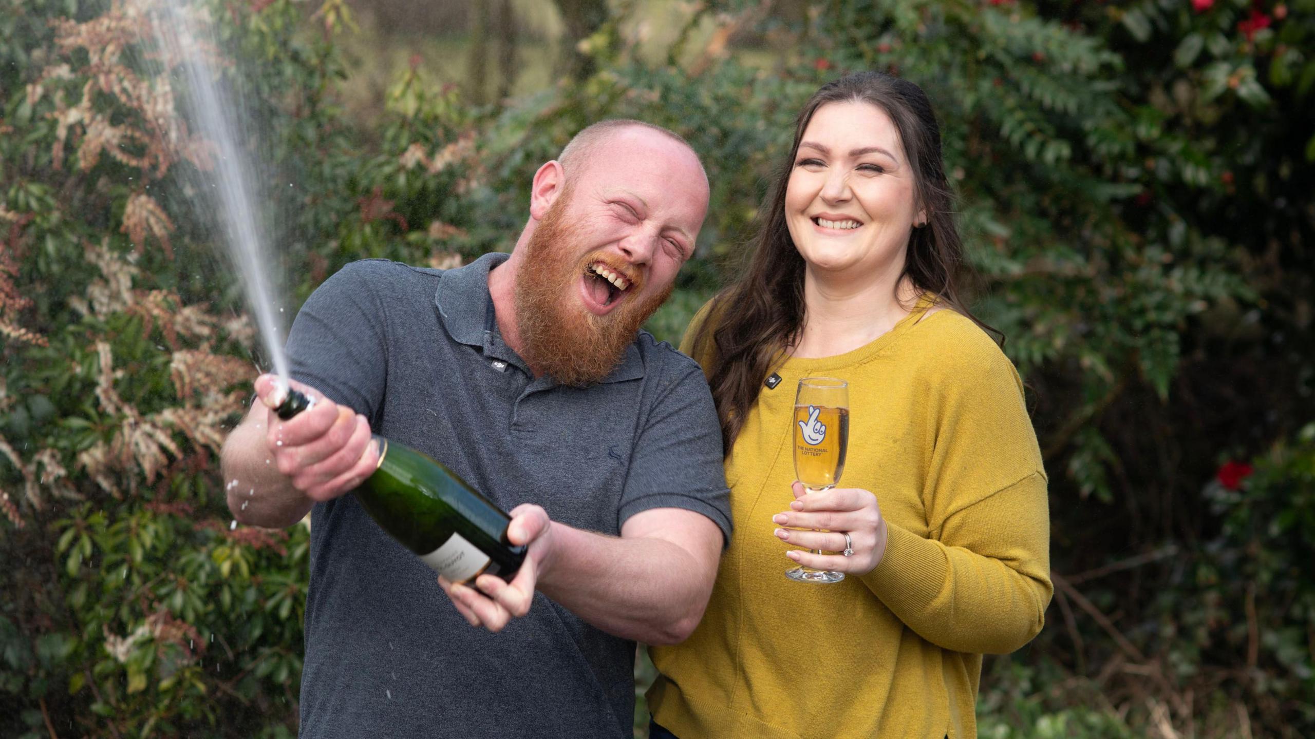 Craig opening a bottle of champagne and spraying it everywhere whilst smiling, and Zoe holding a glass of champagne and also smiling.