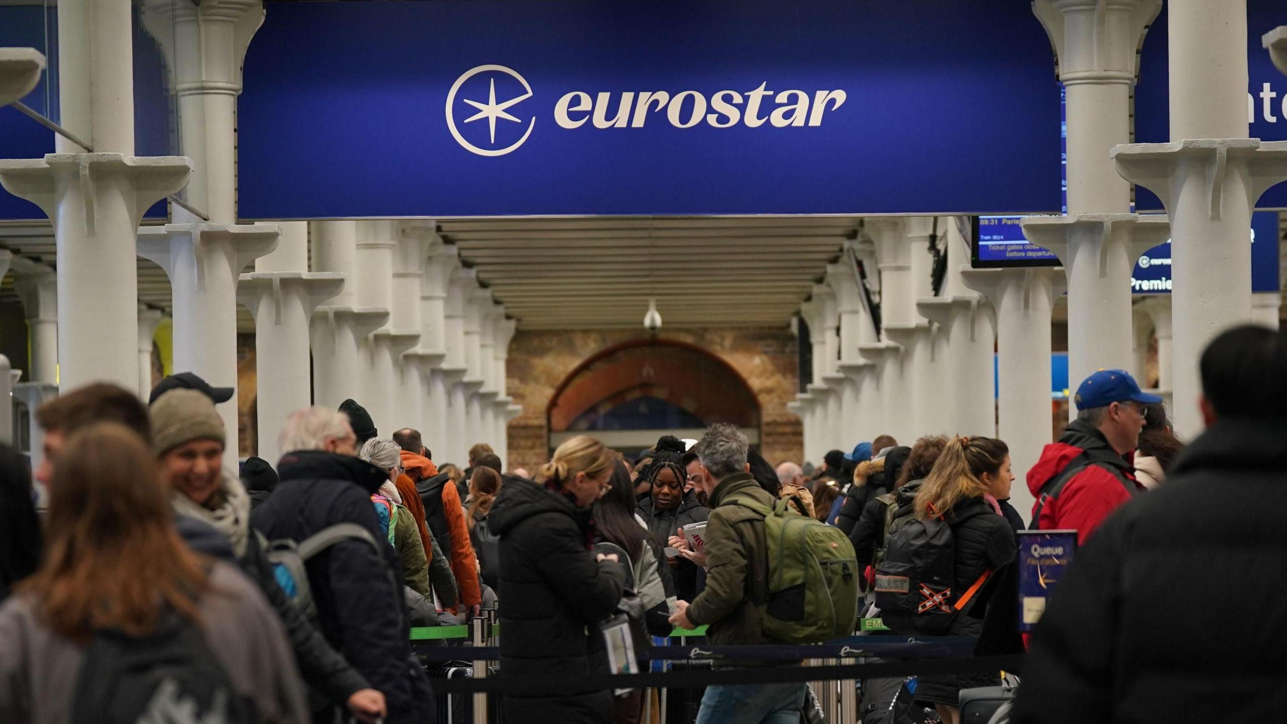 A busy train station, with a blue sign reading "Eurostar" in white letters. There are rows of white pillars the people are queueing in-between and a lot of the people are wearing backpacks.