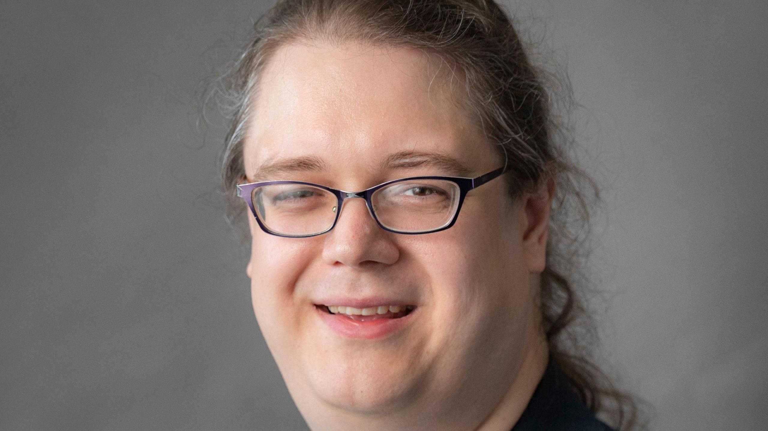 Headshot of Chris Hilliard - they are wearing a black jacket and black rimmed glasses with long hair in a ponytail