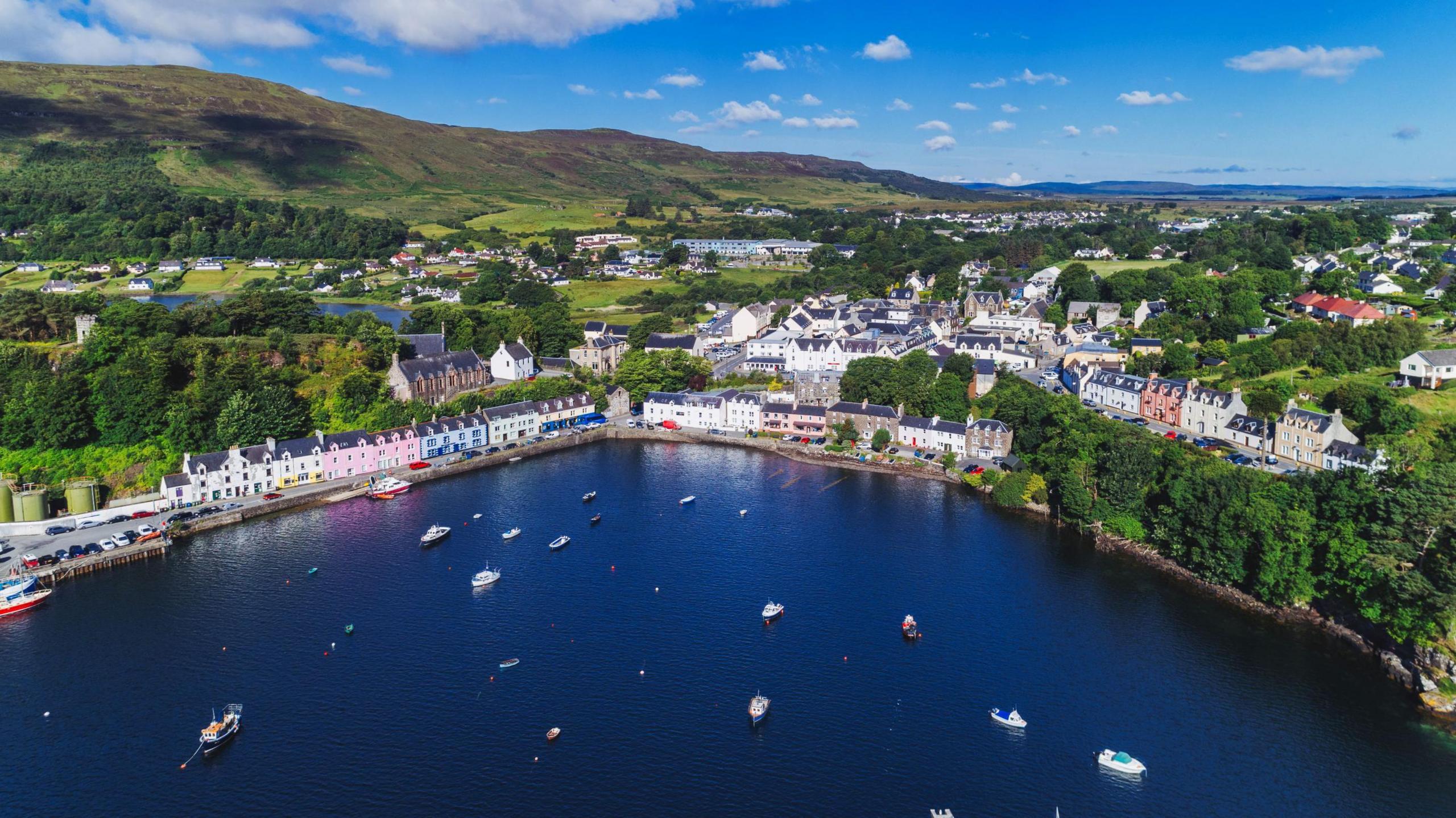 An aerial view of Portree on the Isle of Skye showing brightly coloured houses on the harbourside and boats in the bay. It is a sunny day with fluffy white clouds in the sky.