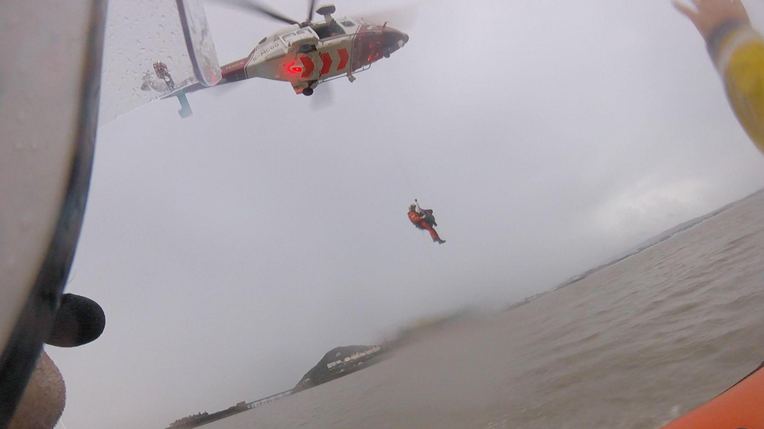 A coast guard crew member is winching Mr Grant into the helicopter to safely transport him to hospital. The scene is seen from the life boat. The water and sky are both grey.