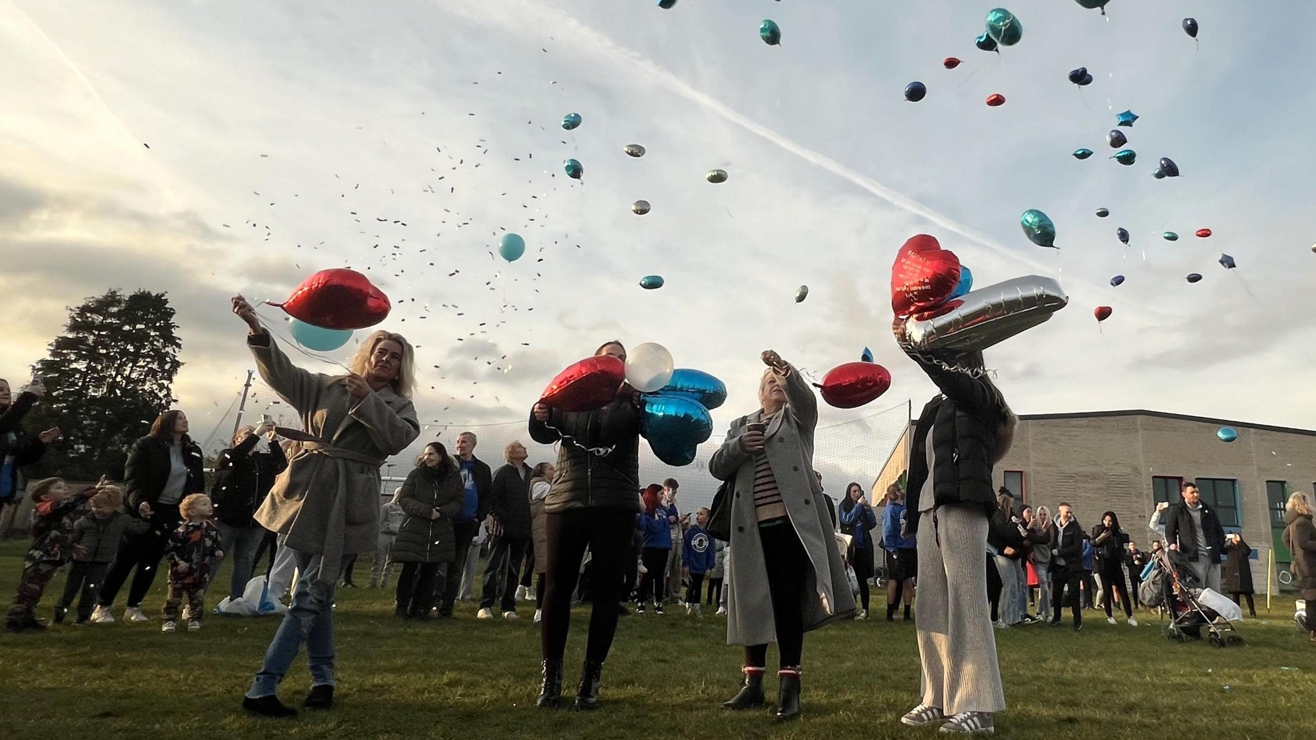 Four women stand on an area of parkland in Bristol releasing balloons. Behind them are many more people also releasing balloons of different colours