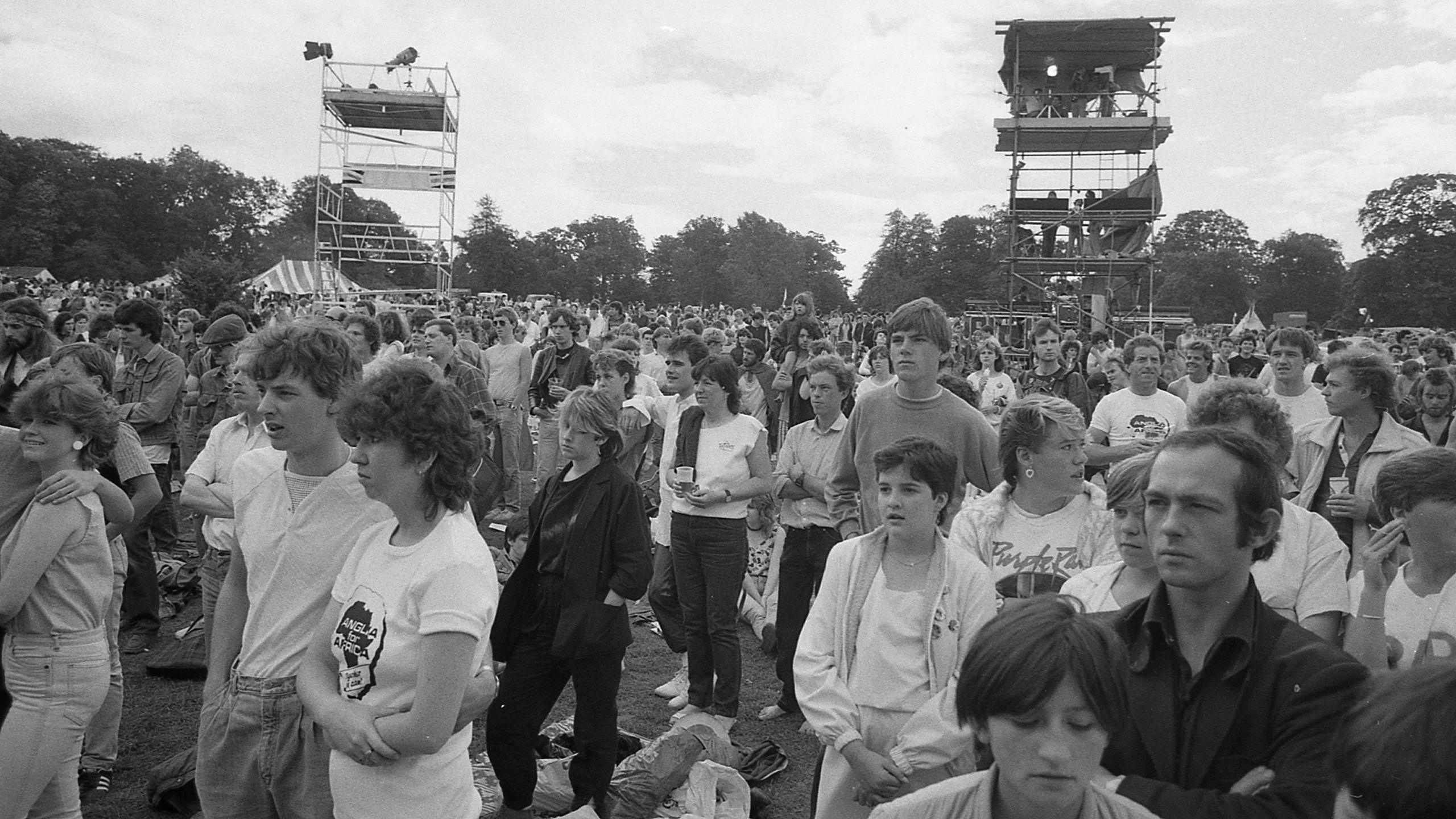 A black-and-white shot of a large crowd of people, mostly young in their teens and 20s, with 1980s clothing and hairstyles. It is an outdoor concert in daylight, and they are all looking towards the stage which is out of shot to the left of frame. Behind them in the background are two scaffolding towers, and a line of trees.