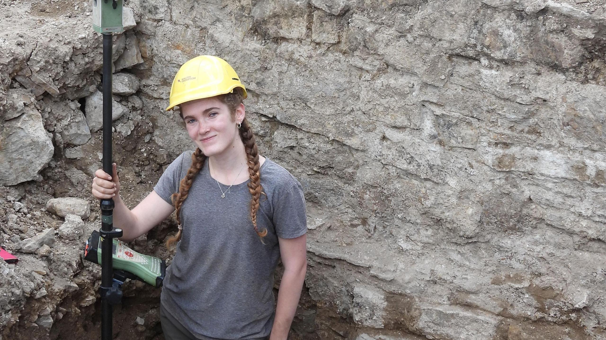 Frankie Rickett, in a grey t-shirt and a yellow helmet, is standing in trench in front of the medieval wall. Her ginger hair is in two braids.
