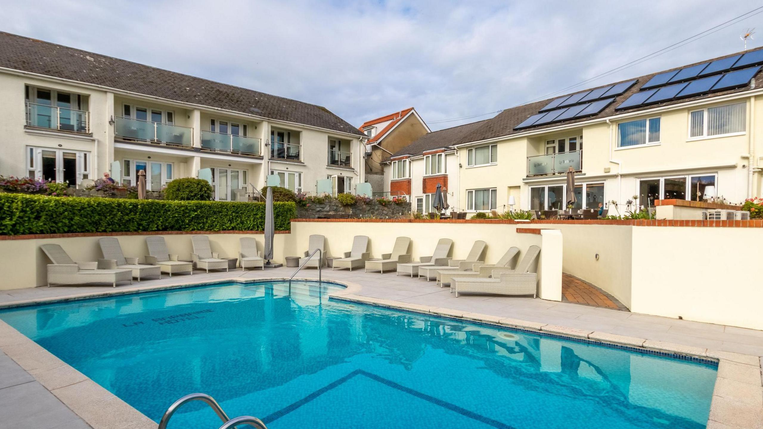 A bright blue swimming pool, surrounded by white sun loungers and white buildings with balconies and solar panels.