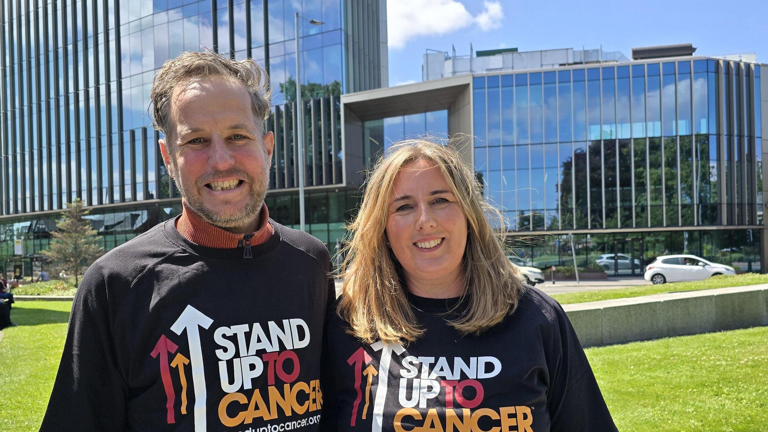 Alex, who has brown hair and beard and Emma Davies, with long blond hair, standing outside the entrance to Manchester Cancer Research Centre wearing t-shirts which read Stand Up To Cancer. They are both smiling. It is a sunny day.