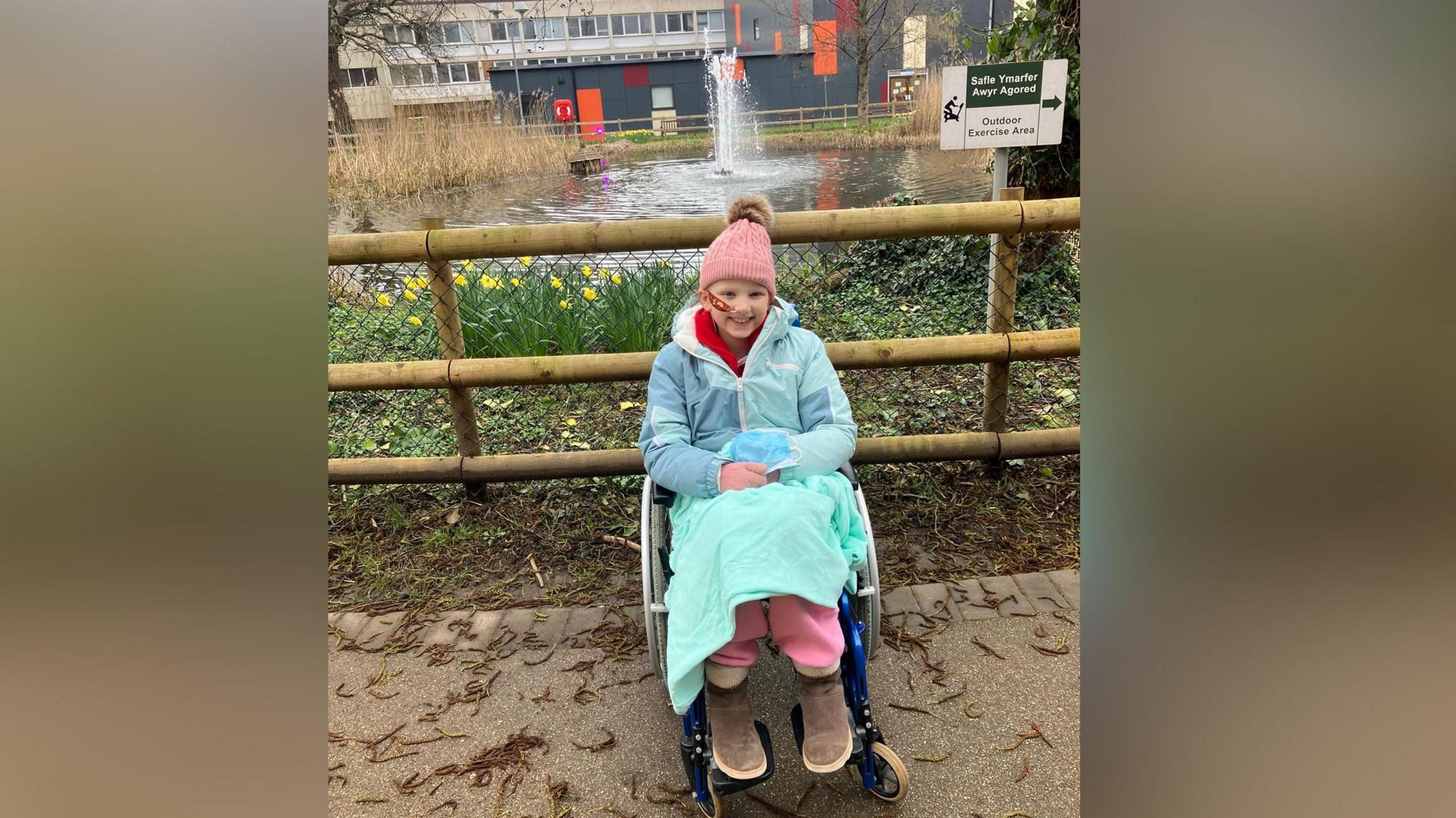 Llun o Kathryn yn eistedd yng nghadair olwyn o flaen llyn. Mae'n gwenu arth edrych ar y camera. Mae'n gwisgo het binc, cot las, menig pinc, trowsus pinc ac esgidiau brown. Mae ganddi flanced dros ei choesau hefyd.