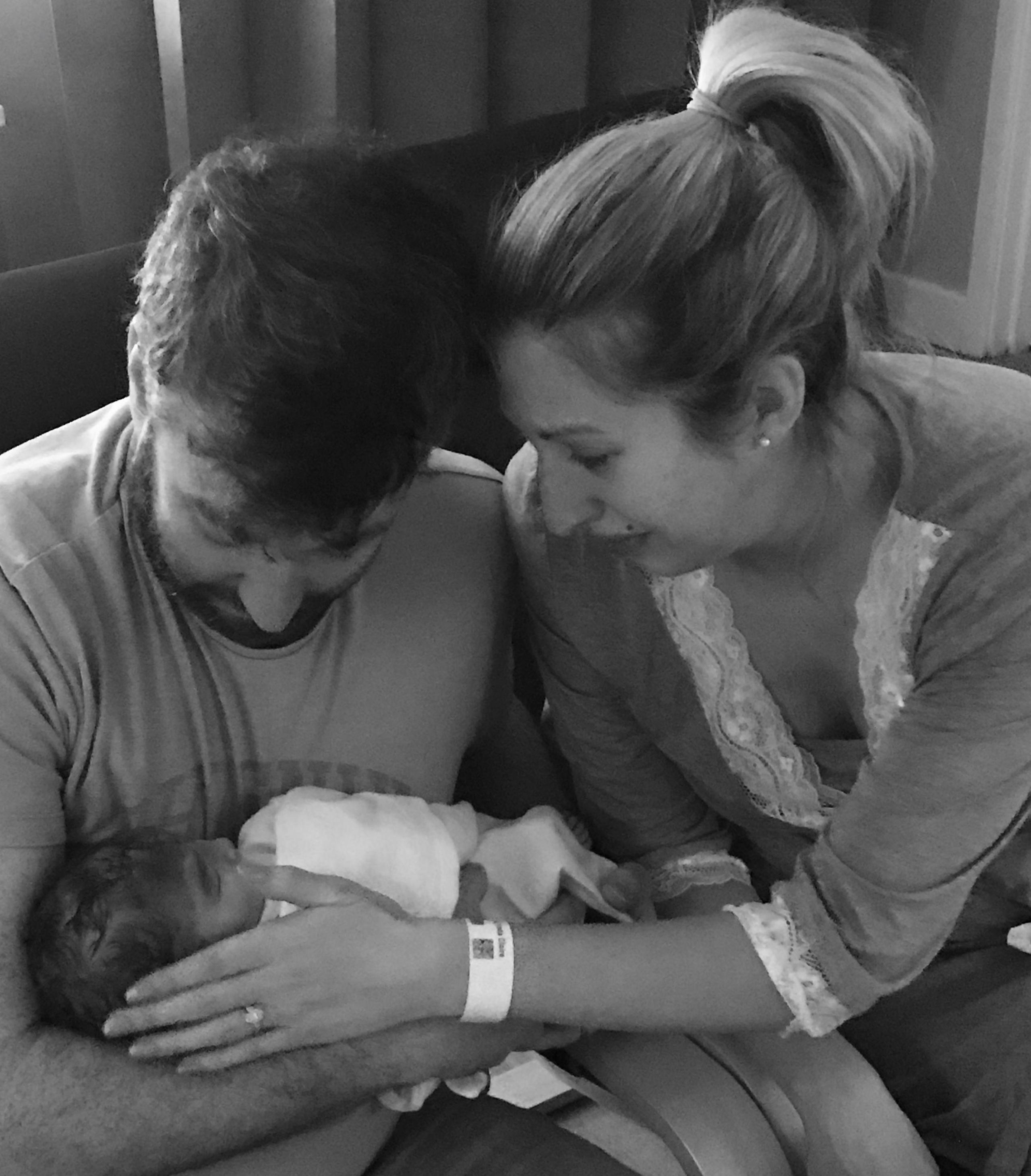 Black and white photo of Dan and Fiona cradling their newborn baby. Dan holds her in his arms and is looking down at her, while Fiona strokes her face. A hospital band is visible on her wrist. 