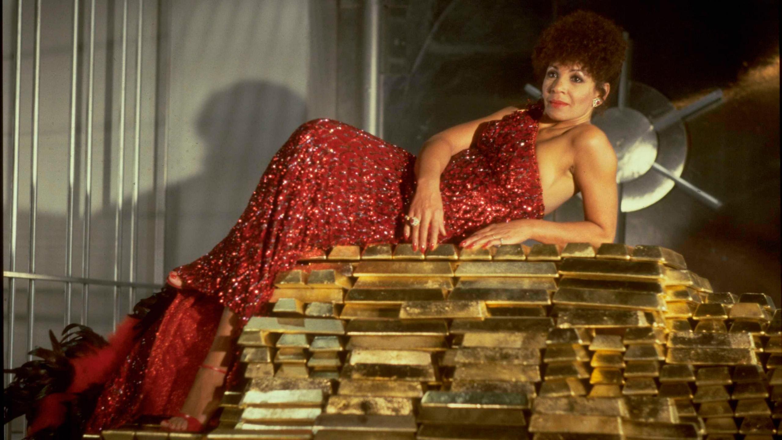 Shirley Bassey lays atop a stack of gold bars