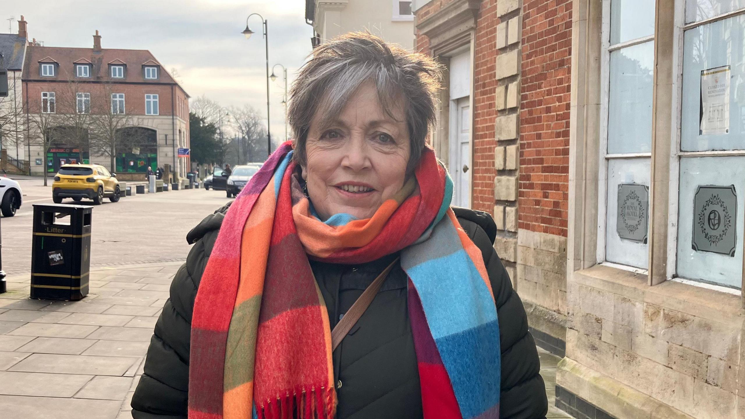 Ally Gosling smiles at the camera as she is photographed standing on a pavement in a town centre. She is wearing a large multi-coloured scarf, a green puffer jacket and a brown cross-body bag. She has short grey hair. Behind her is a yellow car driving down the road. 