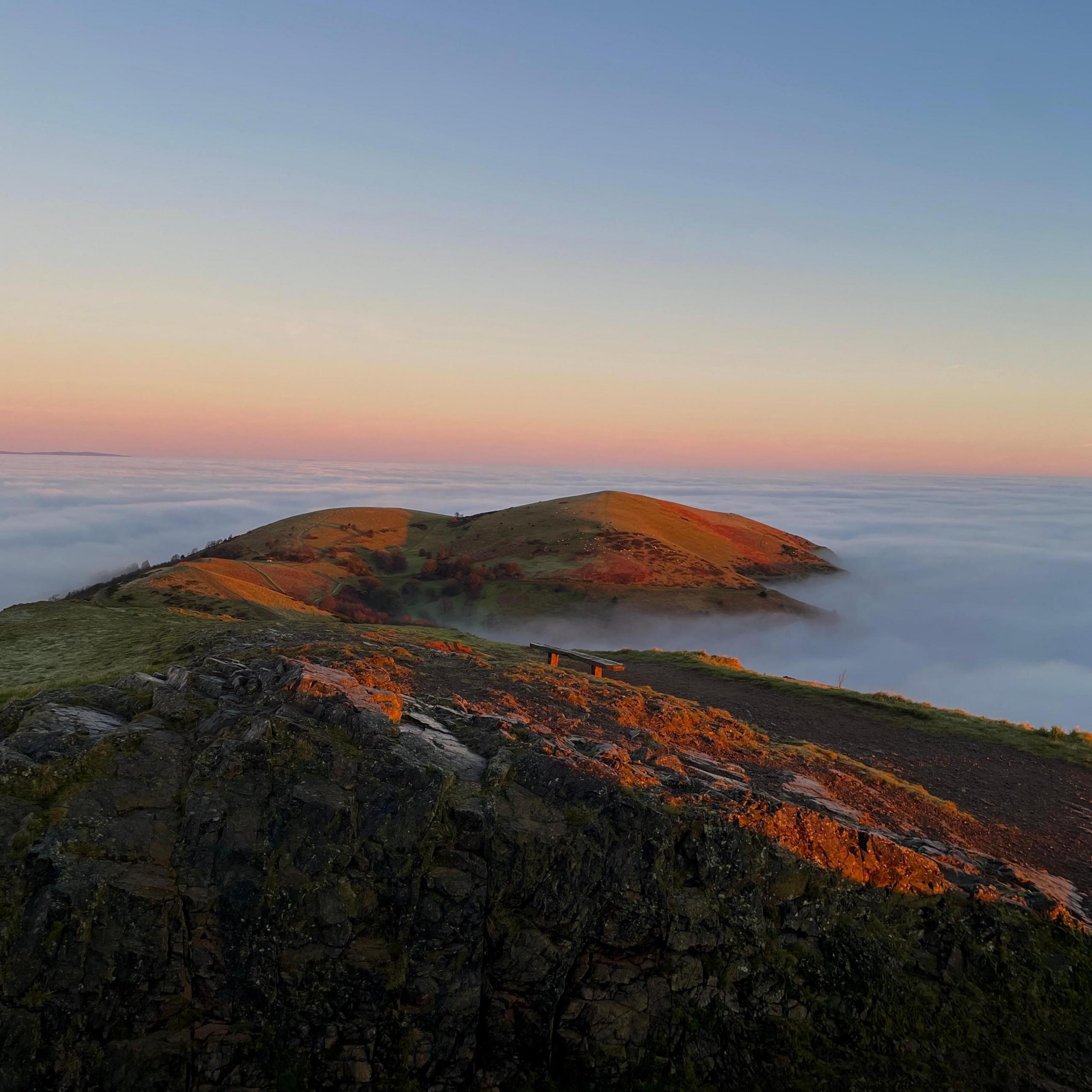 A hill at sunset, with pinks and reds shining on the grass and rocks. Low cloud or heavy mist gives the impression of waves lapping at the foot of the hill.