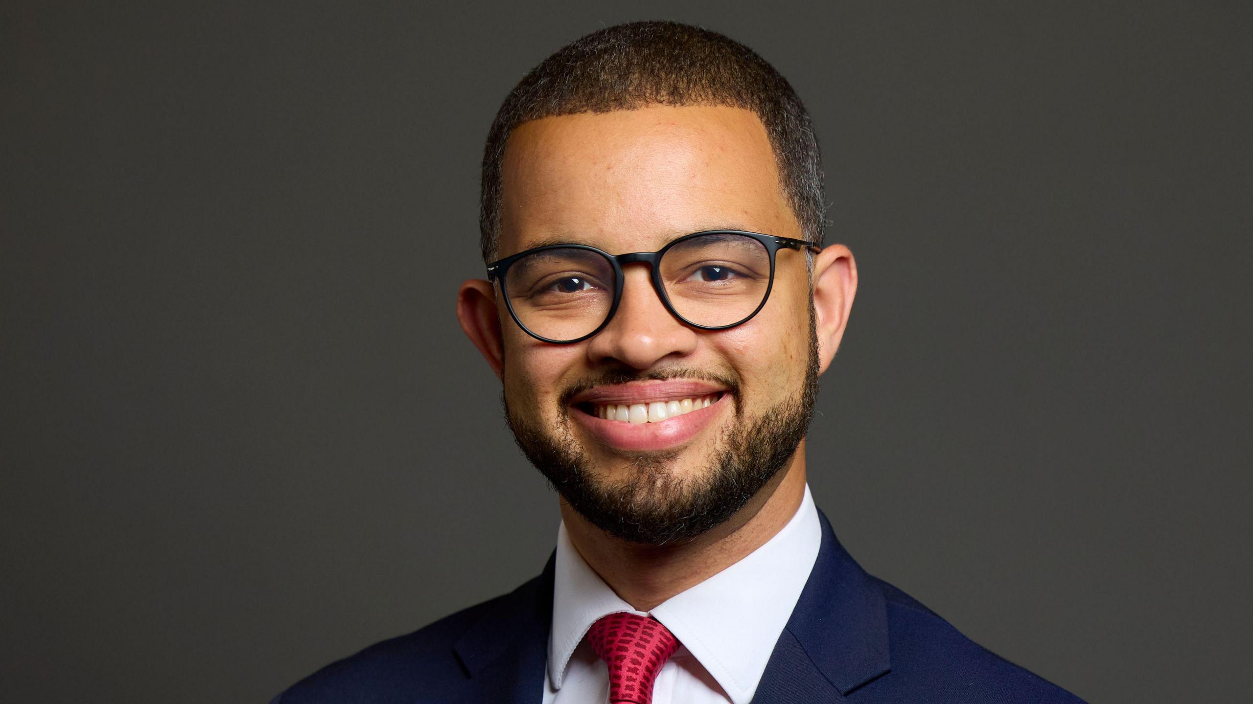 A man with glasses smiling while he wears a suit with a red tie.