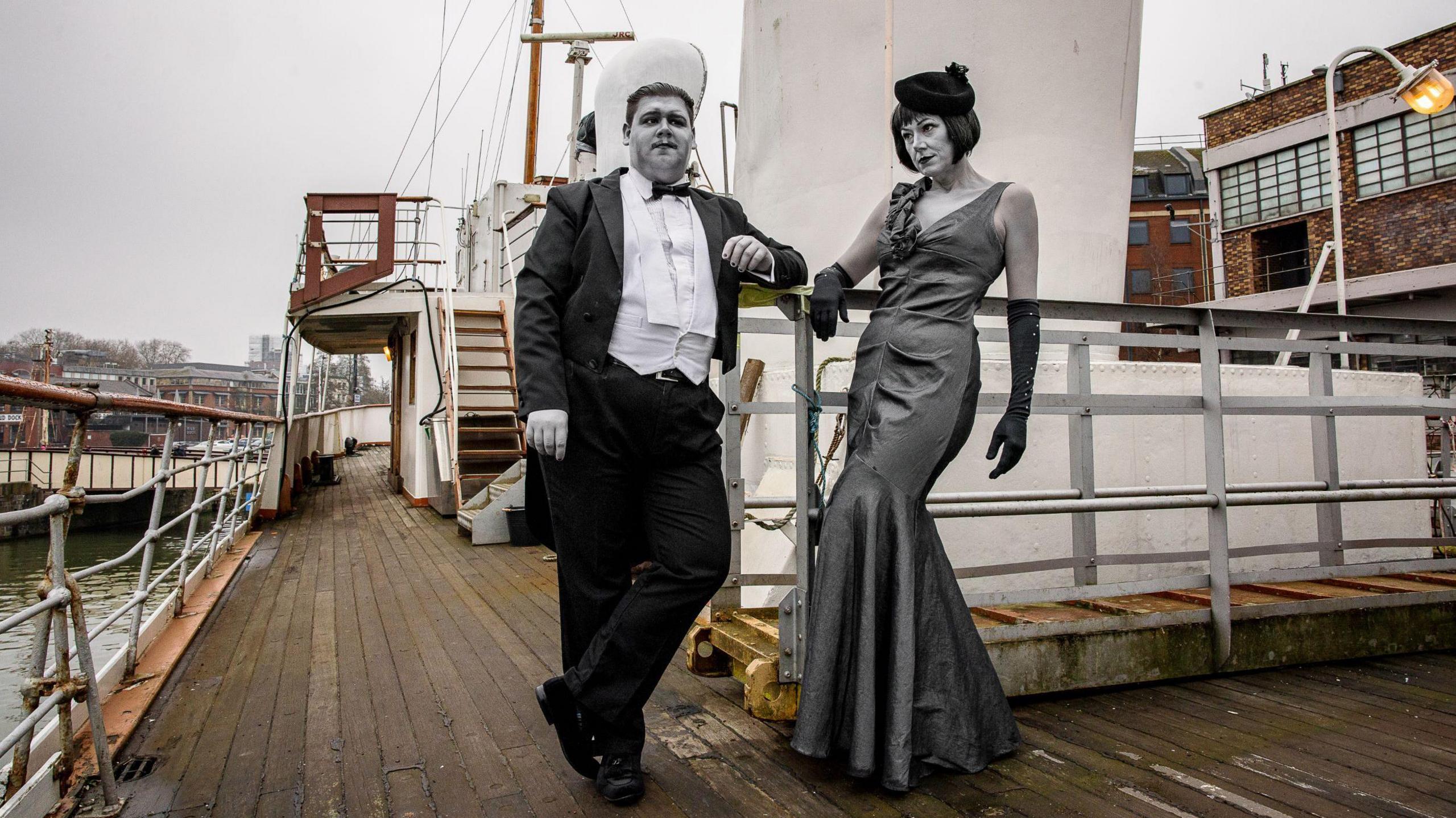 A male actor wearing a tuxedo, white shirt and bow tie, and a female actor wearing a long ballgown stand on a ship's deck in Bristol. It is a colour image but the actors appear as though they are in black and white, thanks to their clothing and extensive make-up. The image is promoting the 2025 Bristol Slapstick Festival