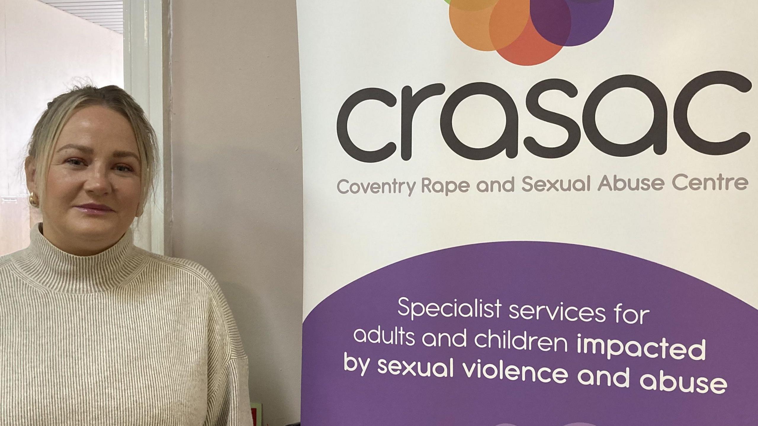 Laura is wearing a cream coloured jumper and is standing in front of a CRASAC promotional stand