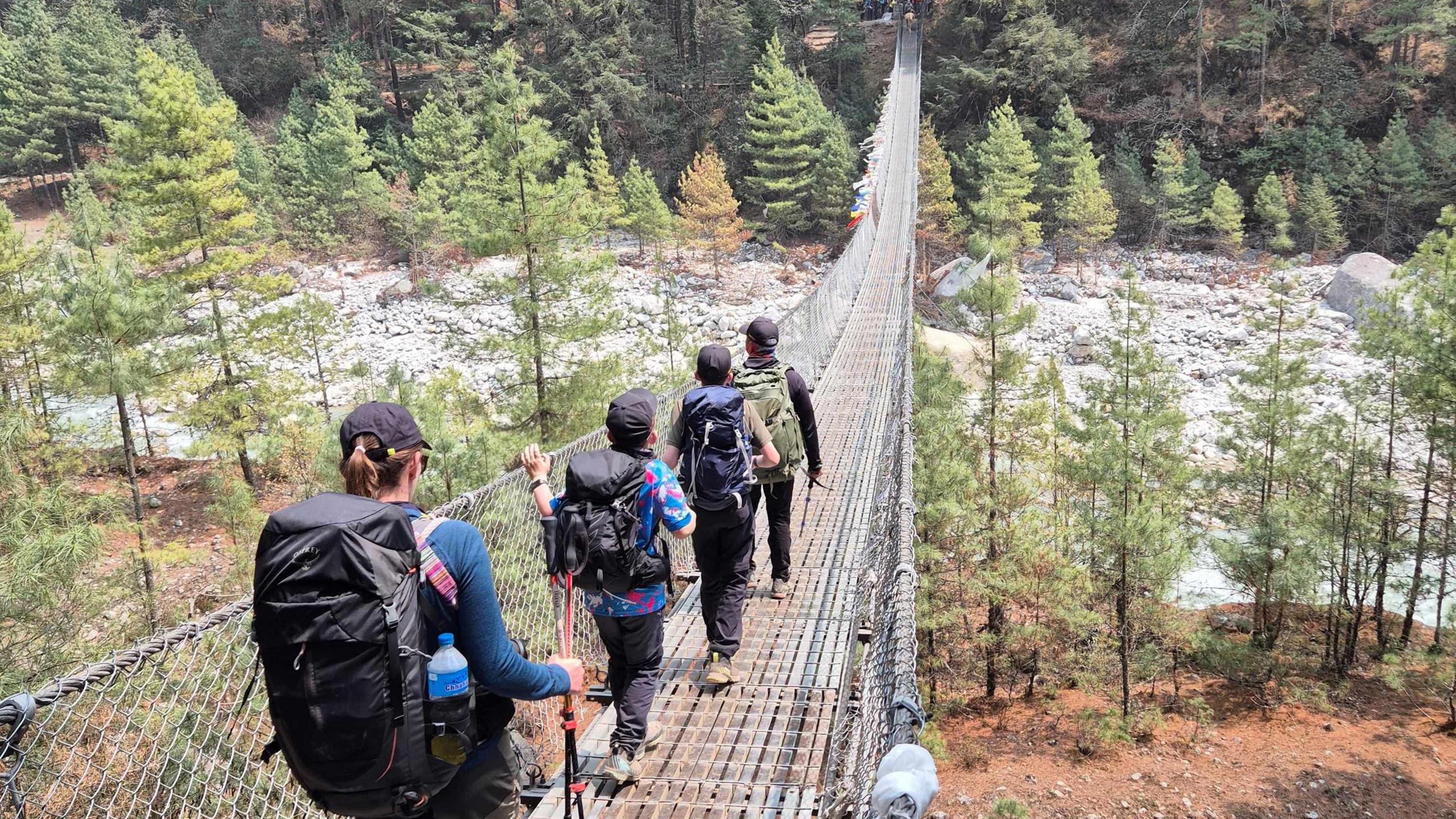 A family of four walking along a suspension bridge into a forest. Mr Russell-Smith at the front followed by two young sons and Mrs Russell- Smith at the back. All four family members have sports wear on,  holding walking sticks and wearing backpacks.