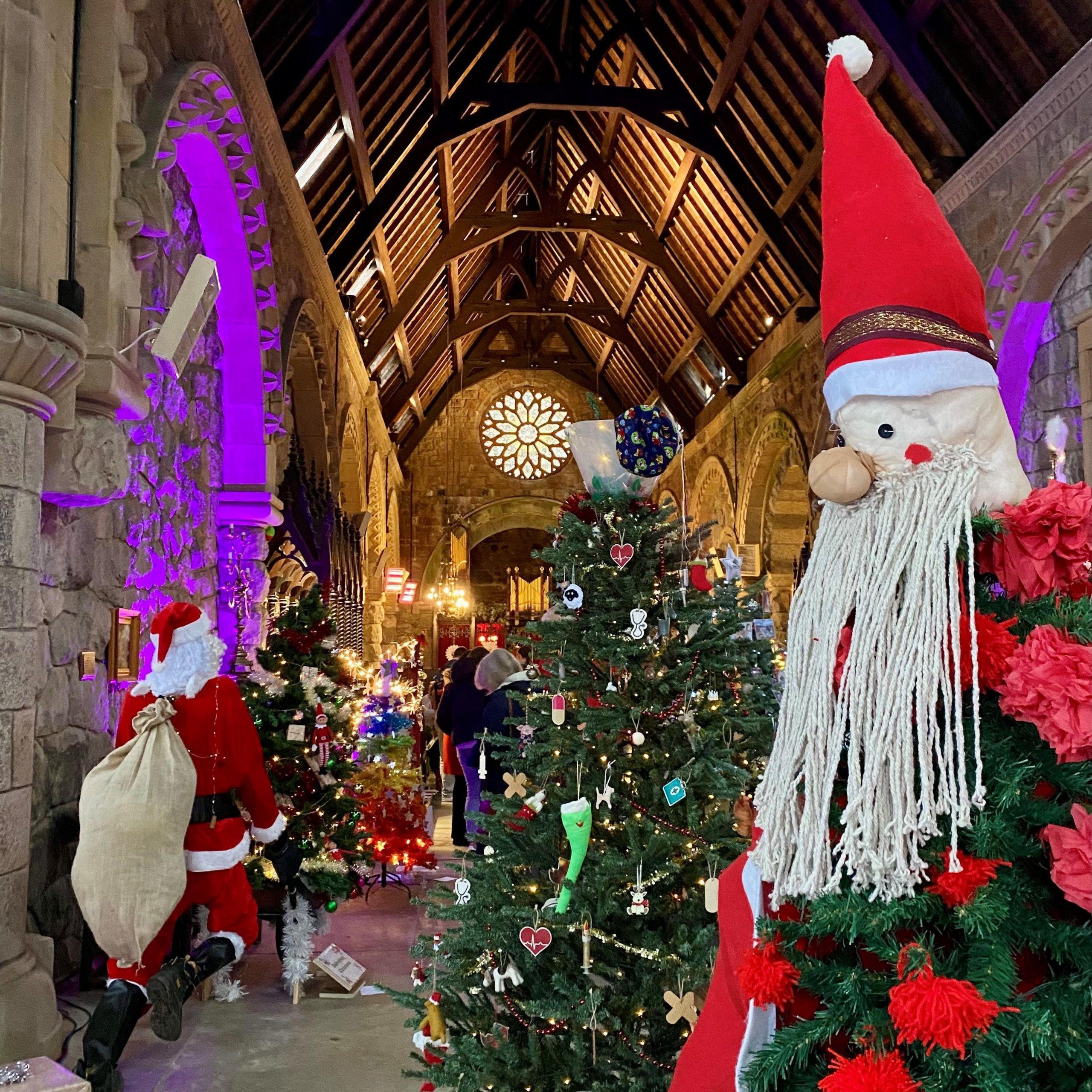 A Santa with a long string beard is on the right of the picture. On the left you can see another Santa with a sack. He has his back to the camera. They are large stuffed toys. The ceiling is wooden and there is a Christmas tree in the centre of the picture.