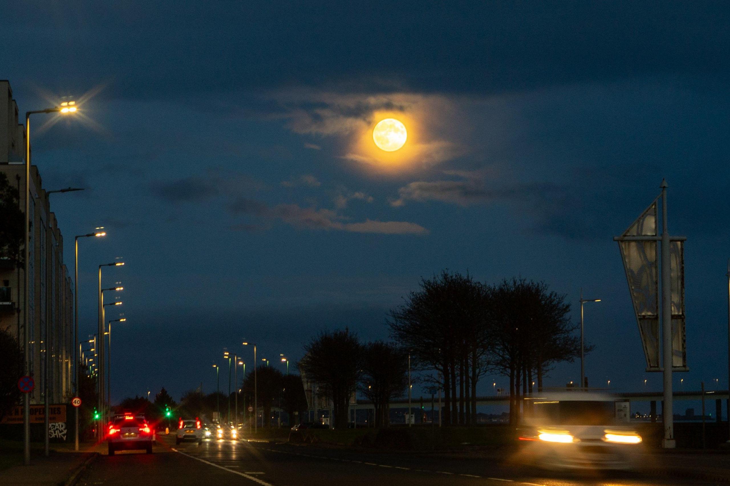 A very bright moon can be seen in the night sky above a road lined with trees and buildings. Cars travelling along the road are blurred.