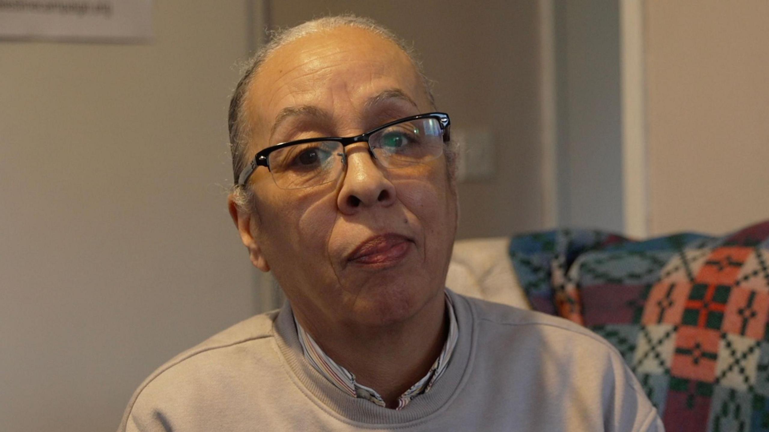 Head and shoulders shot of Winnie Watson who is wearing glasses, has grey tied back hair and is wearing a grey jumper while sat on a safa covered in a tartan rug.