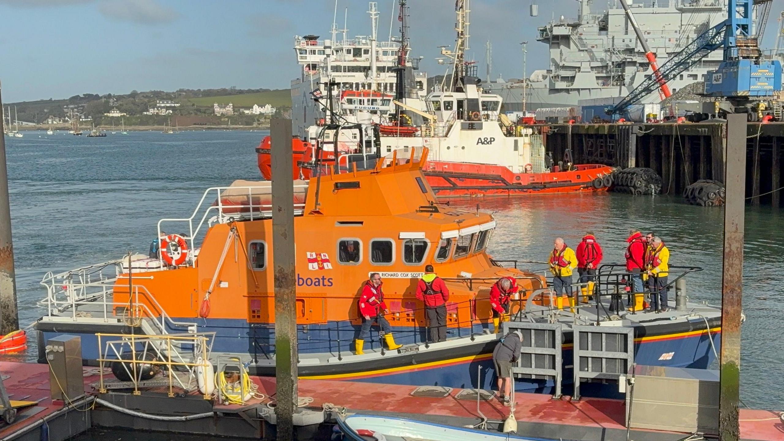 RNLI lifeboat crews dressed in red or yellow jackets and life-jackets prepare the distinctive orange lifeboat for her final voyage from Falmouth. It is a bright sunny day and there is a ferry and a naval vessel on the sea in the background.