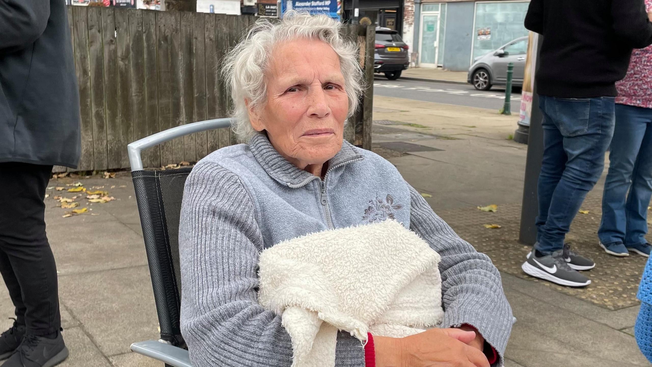 June Gretton, 90, is sat in a camping chair outdoors. A fluffy blanket is covering her legs and torso.