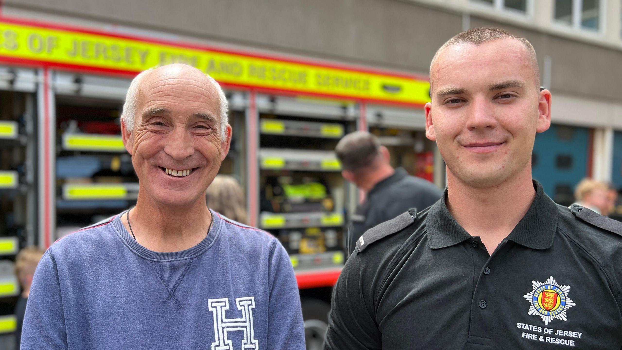 Roger and Logan smile at the camera as they stand in front of a fire engine which is being shown off to the public. Roger is on the left and as a blue jumper on with a white letter H on his chest. Logan is in a firefighter polo T-shirt.