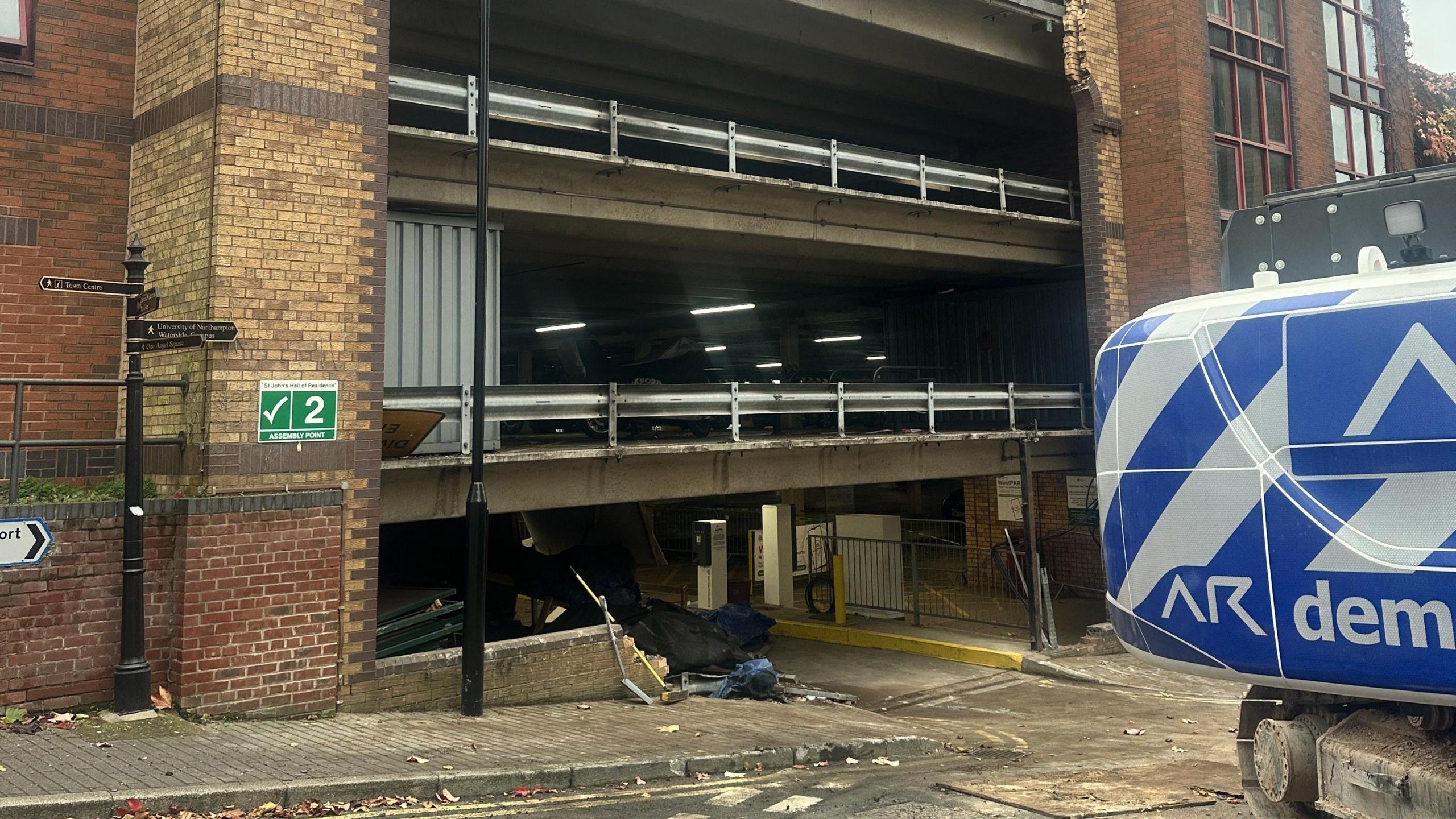 Damage to St John's car park. The entrance to the car park is scarred by broken brickwork and a piece of machinery with an implied purpose in securing the site is partially pictured.