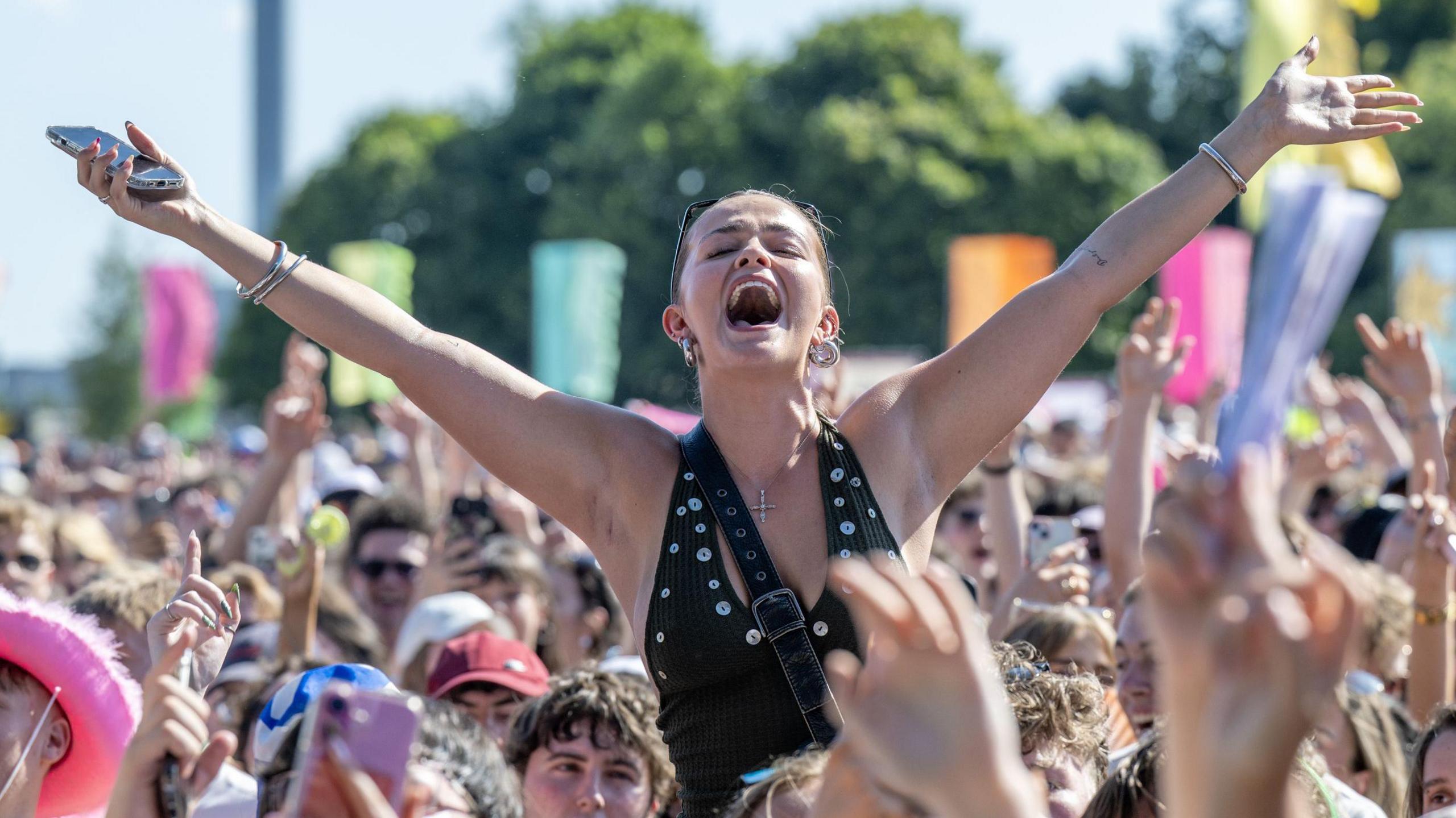 A crowd shot of TRNSMT. One woman is on someone's shoulders and has her hands in the air. People in the crowd have their phones up or their arms in the air. 