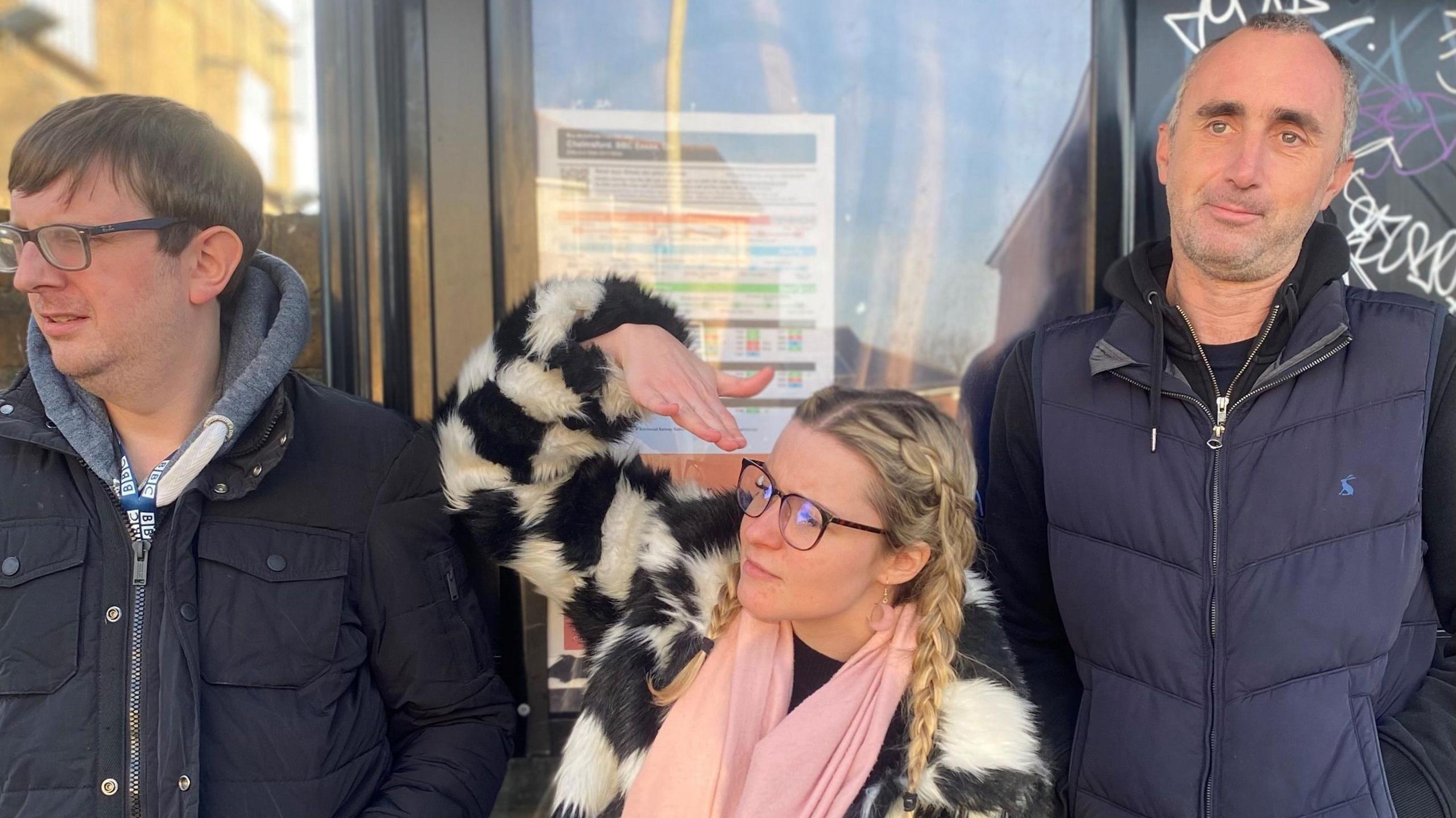 Adam Bennett, Robyn Wallis and Ben Fryer standing at a bus stop. Bennett is wearing a black coat and glasses. He has brown hair. Wallis is wearing a black and white furry coat. She is wearing glasses and has plaited blonde hair. Fryer is wearing a navy gilet over a black hoodie. He has grey hair.