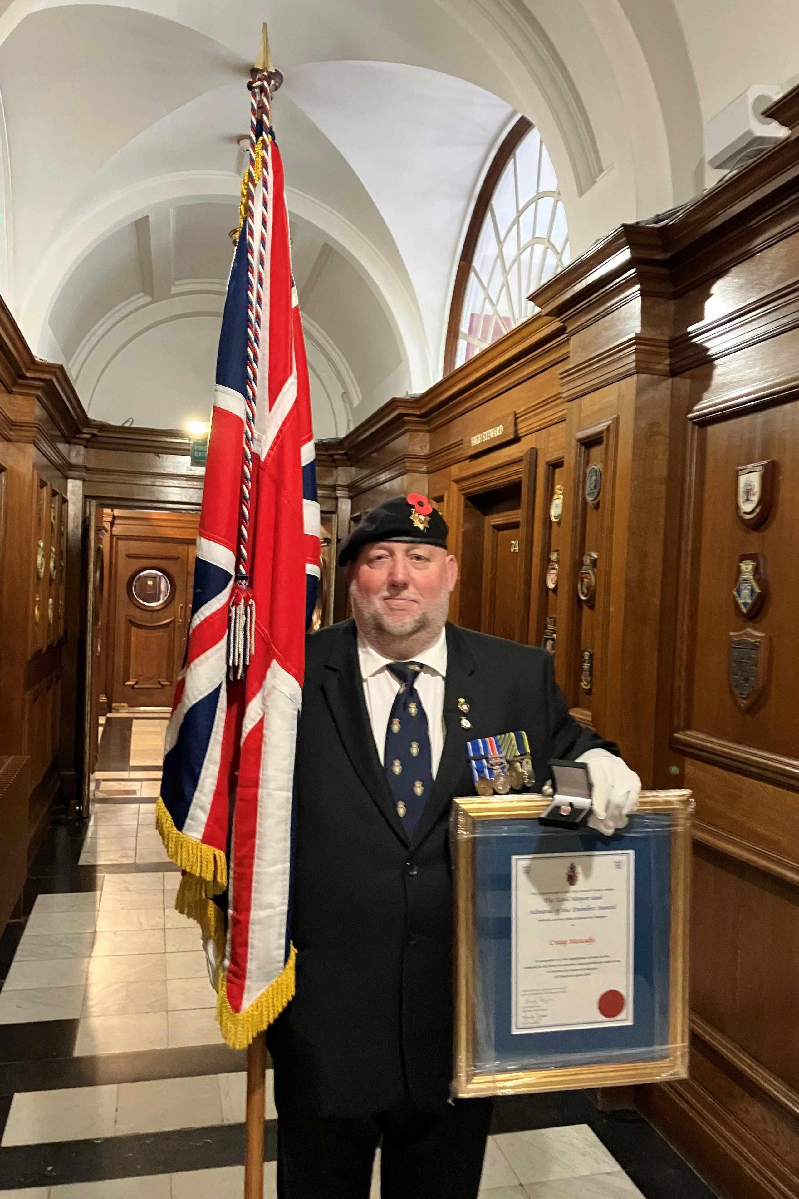 Man in smart blazer and regimental beret and tie holding a medal and a framed certificate in his left hand and a Union flag standard in the other. He is standing in a wood-panelled corridor.