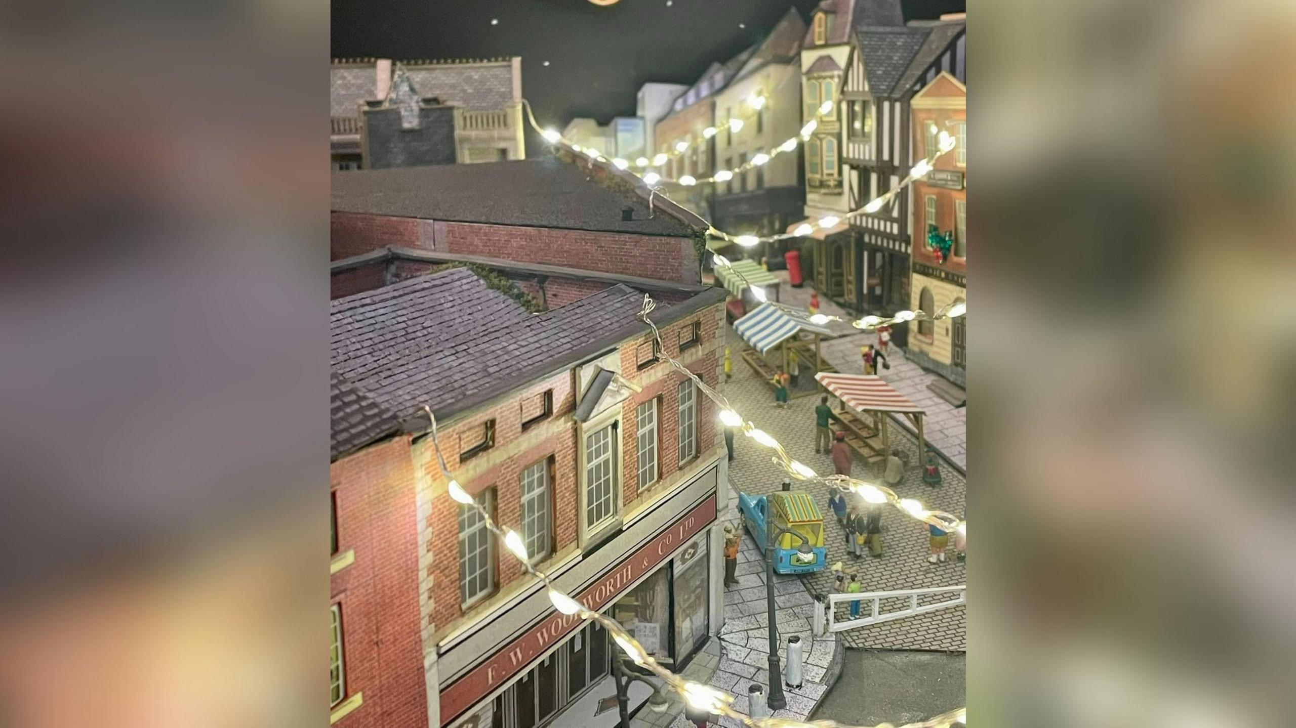 A close up of the model village strung with Christmas lights and showing the bottom part of the cobbled High Street. There are mini market stalls and some buildings are clad in a Tudor style, with a Woolworth's store also seen in the scene.