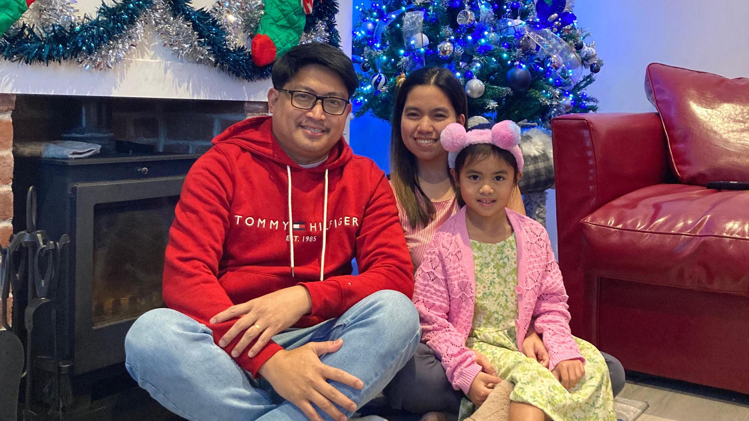 Oliver and Russel Fabia with their daughter. They are sat on the floor in front of a log burner and a Christmas tree.